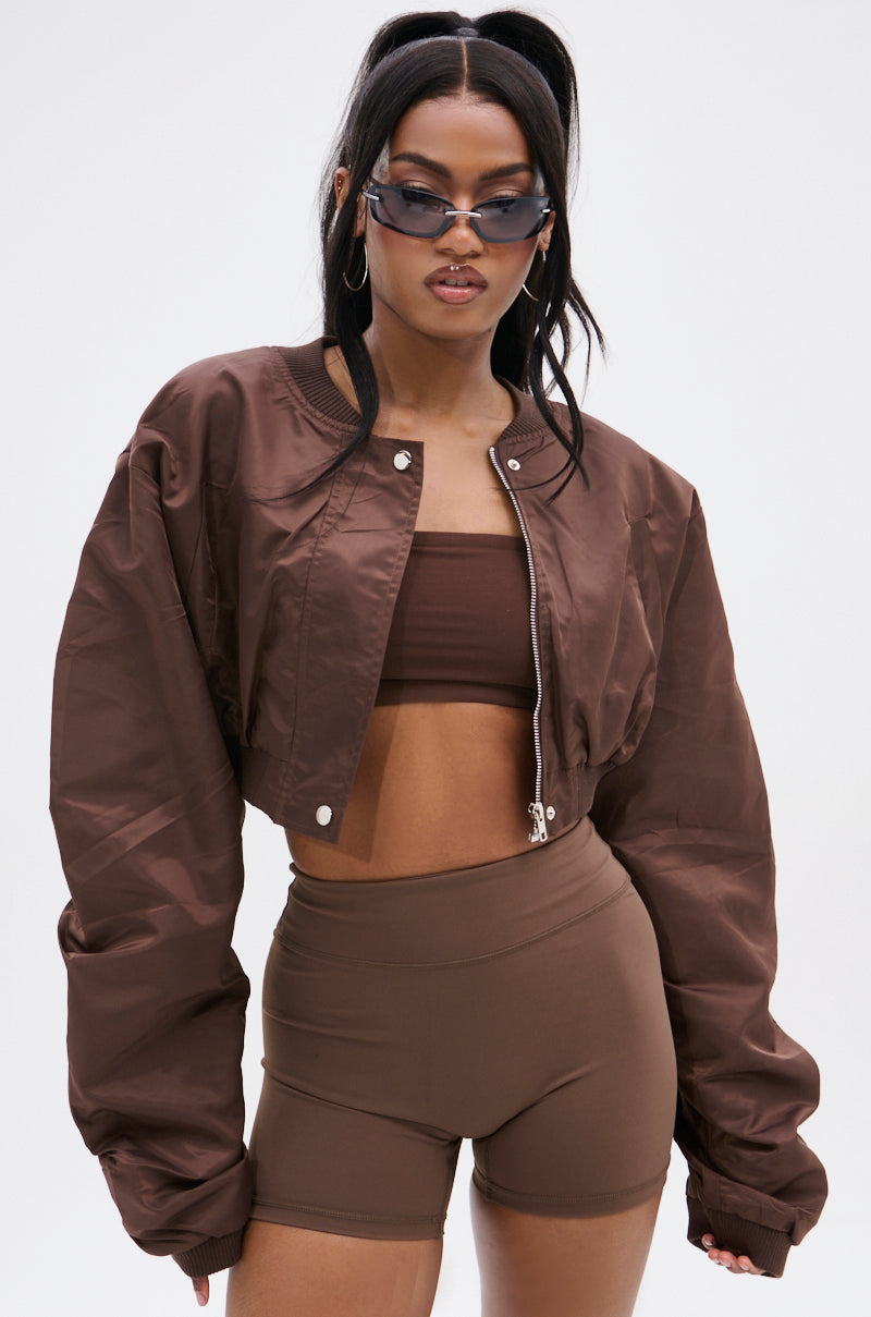 A woman wearing sunglasses, hoop earrings, the TAKE IT EASY CROPPED BOMBER in brown windbreaker fabric, a brown crop top, and high-waisted brown bike shorts stands against a plain white background.
