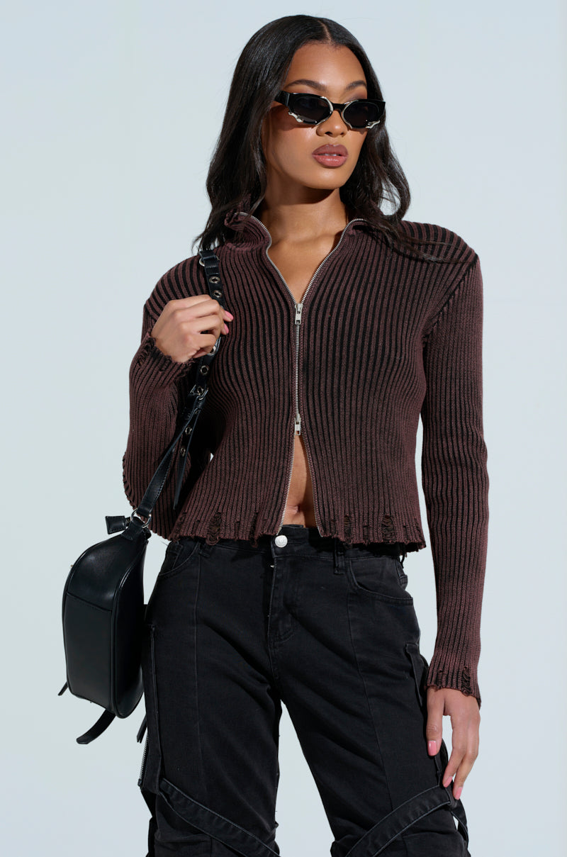 The woman exudes confidence in her AKIRA Label IT'S IN MY NATURE brown ribbed zip-up sweater and black pants, accessorized with sunglasses and a sleek black backpack. She poses against the light background, embodying effortless style.