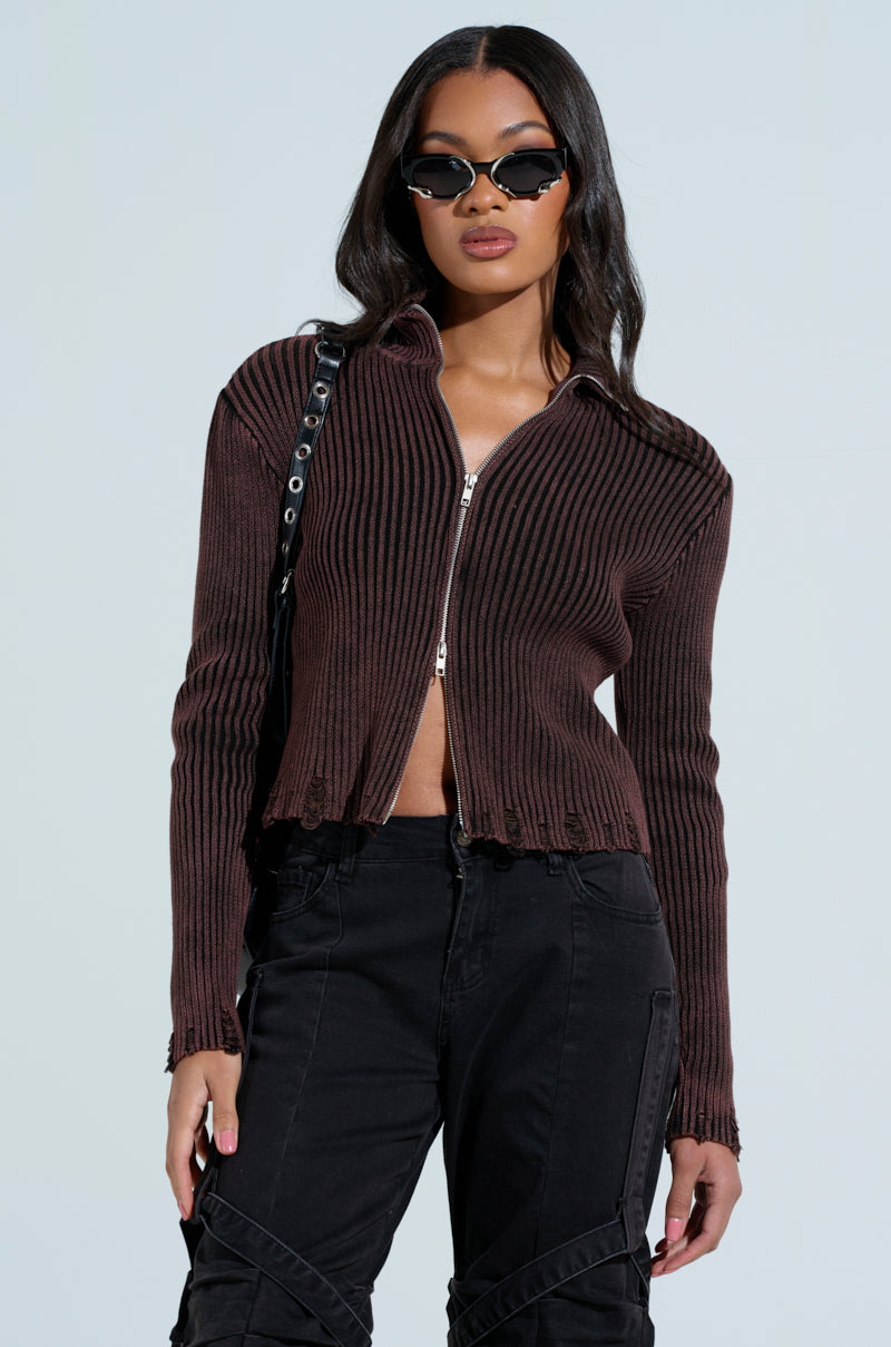 Against a light background, a person exudes confidence in sunglasses, black distressed jeans, and a brown "IT'S IN MY NATURE ZIP UP SWEATER." With one hand slightly raised and a black bag slung over their shoulder, they strike a confident pose.