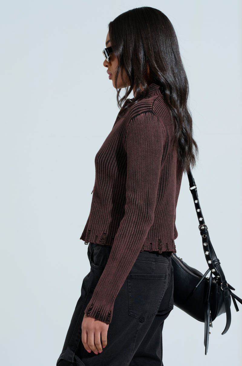 A woman with long dark hair and sunglasses wears a ribbed brown IT'S IN MY NATURE ZIP UP SWEATER. She carries a black bag over her shoulder and dons black distressed jeans while walking against a plain, light-colored background.