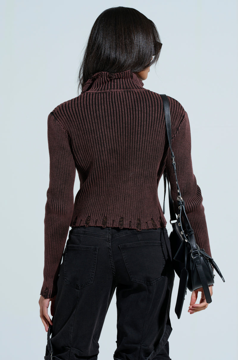 A person with long dark hair wears the AKIRA Label's brown "IT'S IN MY NATURE ZIP UP SWEATER" featuring a ruffled hem and distressed detailing. Paired with black pants, they hold a black shoulder bag, standing turned away from the camera against a light gray background.