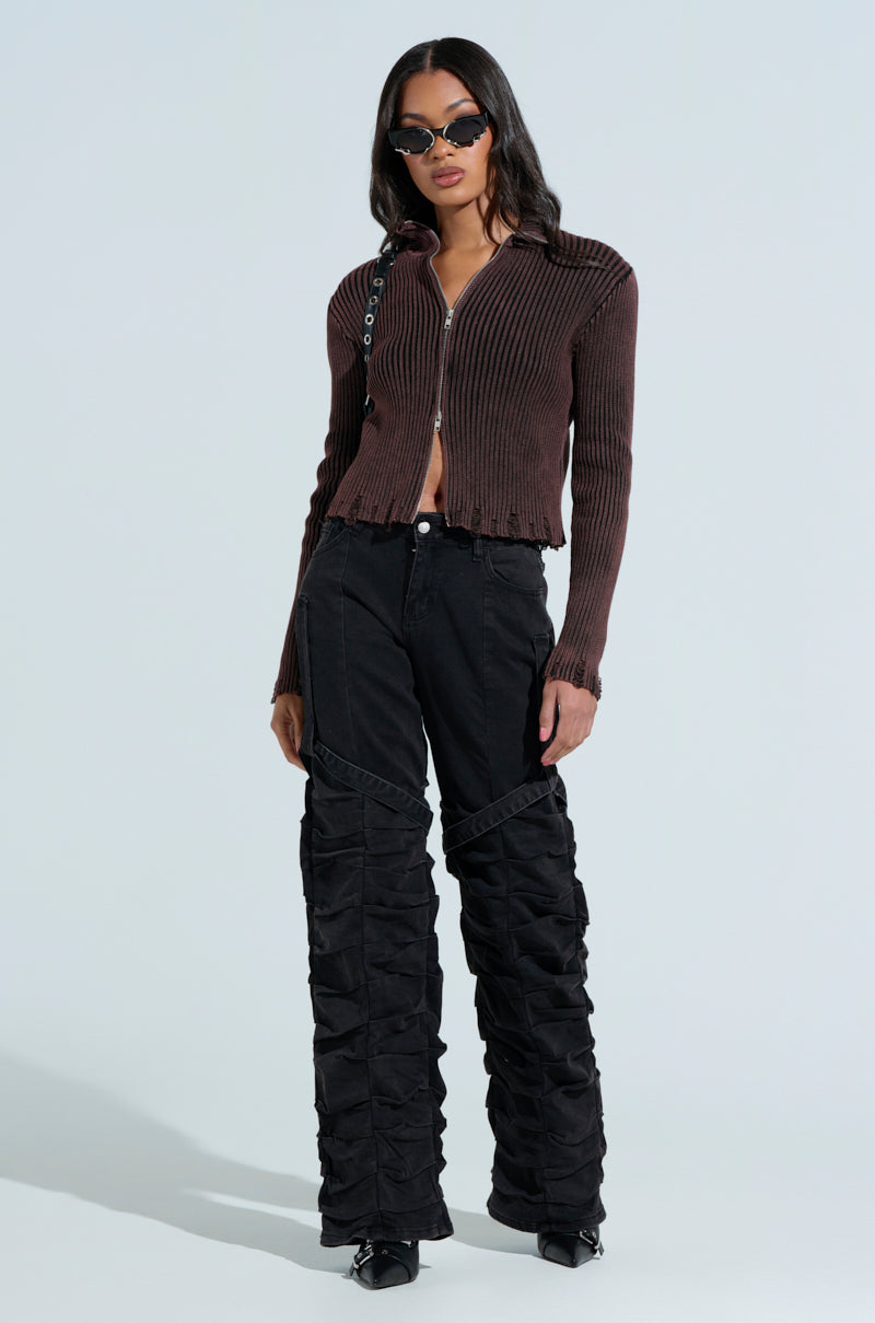 A woman confidently poses in an IT'S IN MY NATURE ZIP UP SWEATER, brown and ribbed, paired with black textured wide-leg pants and pointed shoes. Her long dark hair contrasts with large sunglasses against a plain light background.