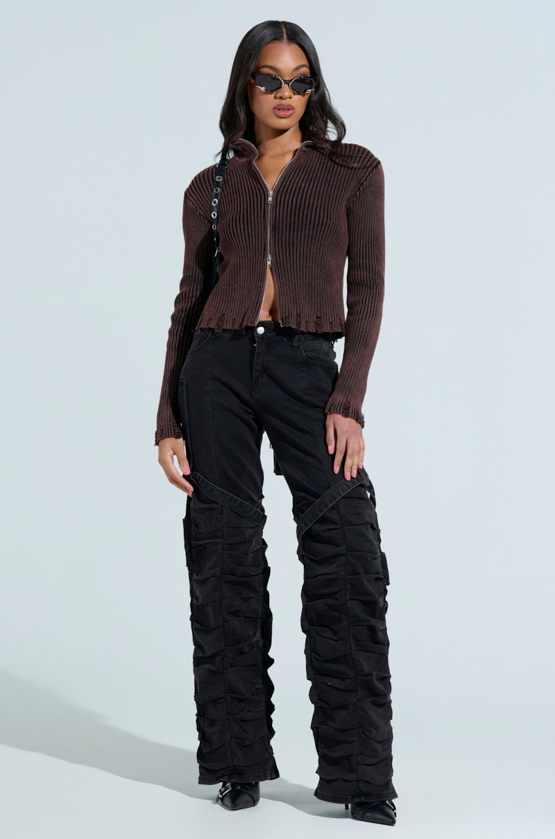A person poses against a plain background wearing the "IT'S IN MY NATURE ZIP UP SWEATER" with black ruched pants and black pointed shoes. With long hair and sunglasses, they carry a bag on one shoulder.