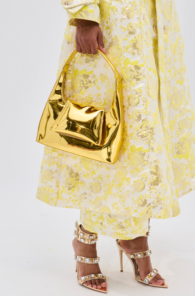 Holding the IN LONDON WITH IT PURSE, a person in a yellow floral dress pairs it with gold-embellished high-heeled sandals for a glamorous, elegant look.