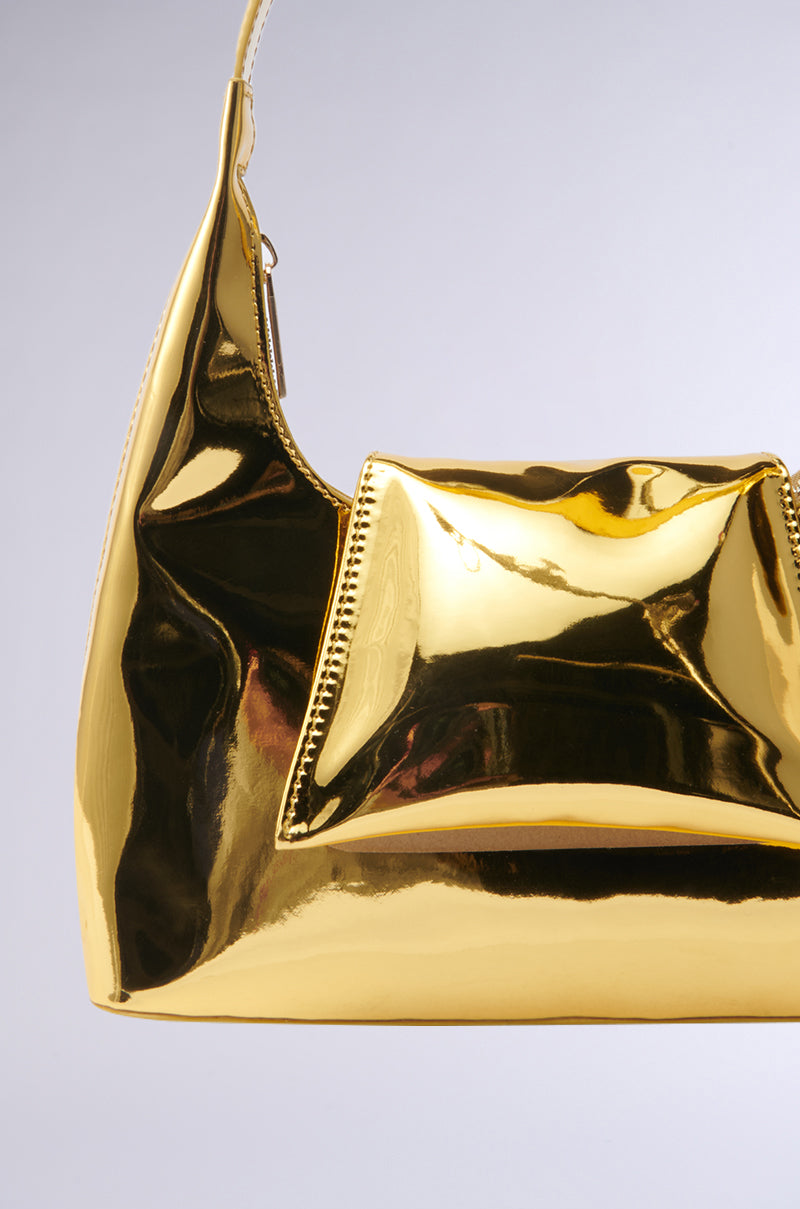 The IN LONDON WITH IT PURSE is a shiny metallic gold shoulder bag with a structured design, smooth reflective surface, and side zipper, making it stand out against a plain light background.