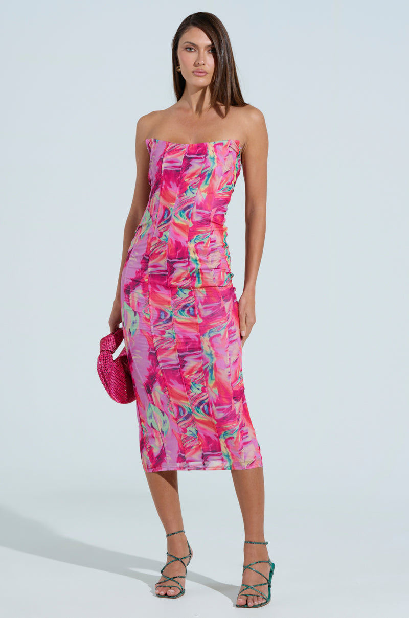 A woman in a strapless, lively print dress poses against a light blue backdrop, holding a bright pink clutch and wearing green strappy heels. Her straight, shoulder-length hair complements the Veronica Printed Mesh Midi Dress's bodycon design perfectly.
