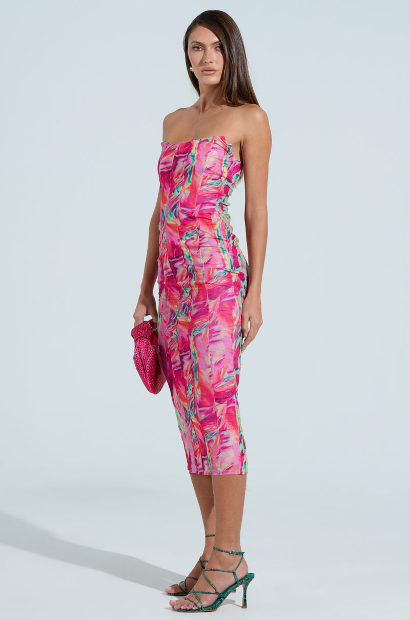 A woman poses in a strapless VERONICA PRINTED MESH MIDI DRESS featuring a vibrant warped print. She holds a textured pink clutch and wears green strappy sandals against a plain light blue background.