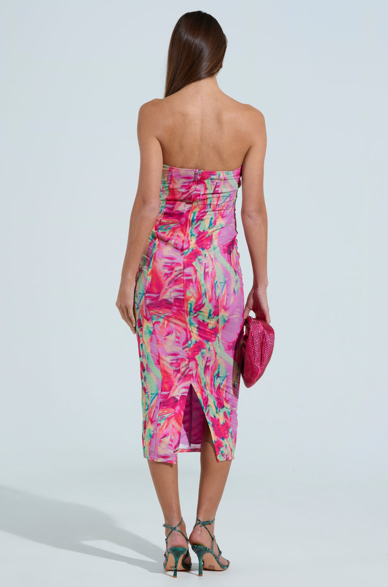 A woman with long hair stands facing away, showcasing the vibrant VERONICA PRINTED MESH MIDI DRESS. She holds a textured pink clutch that complements her teal strappy heels. The bodycon fit of the dress highlights her figure against a plain light background.