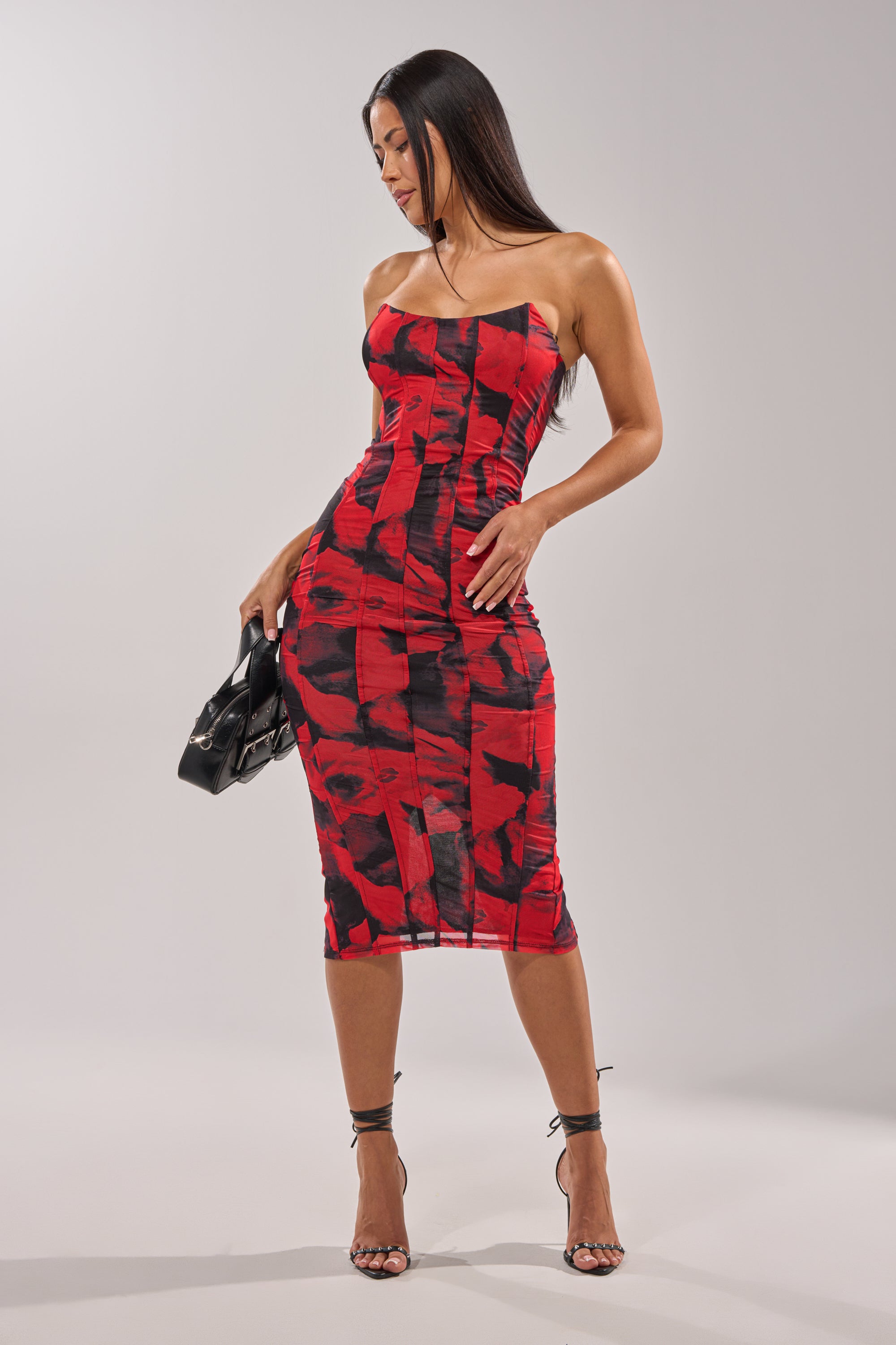 A woman stands against a plain light background wearing the ROSE GARDEN MAXI DRESS, an abstract red and black strapless maxi, paired with a black handbag and black strappy high heels. 0