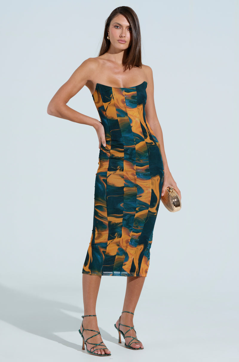 A woman models the SHES UNFORGETTABLE ABSTRACT MESH MIDI DRESS, a strapless bodycon featuring an orange and blue pattern. She pairs it with a small textured clutch and strappy green sandals against a plain light blue backdrop.