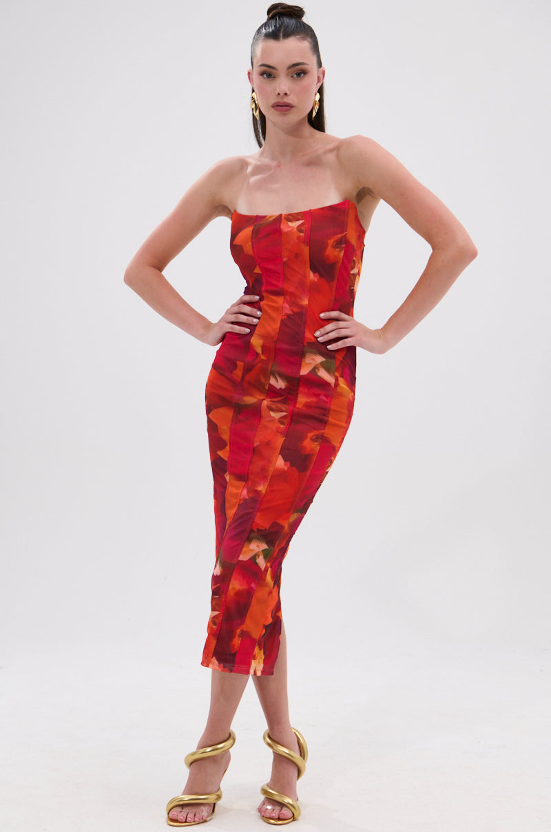 A woman stands against a white background wearing the ARCHETYPE PRINTED MIDI DRESS, a strapless bodycon midi with a bold red and orange abstract print. She pairs it with gold sandals, gold hoop earrings, and a sleek high ponytail. 0