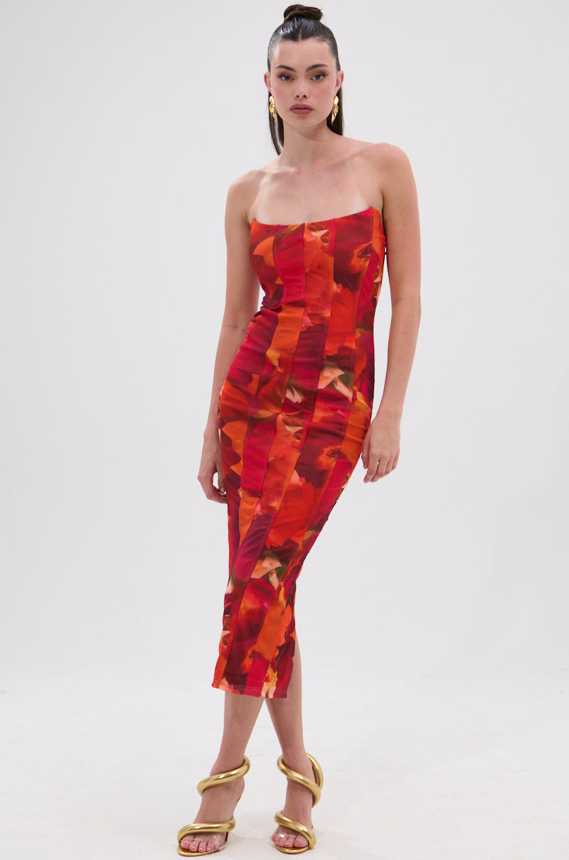 A woman wears the ARCHETYPE PRINTED MIDI DRESS, a strapless bodycon midi with a red and orange abstract print, styled with gold sandals, hoop earrings, and her hair in a high ponytail against a white background. 1