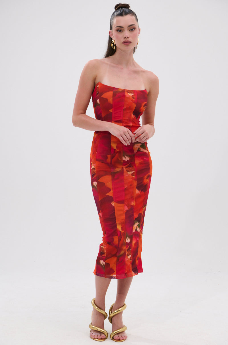 A woman poses against a white background in the ARCHETYPE PRINTED MIDI DRESS, a bodycon midi with a vibrant red and orange abstract print, styled with gold hoop earrings, strappy gold heels, and a high ponytail. 2