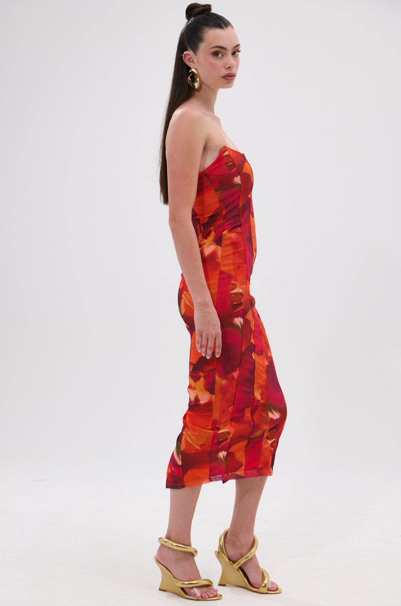 A woman with long dark hair styled half up wears the ARCHETYPE PRINTED MIDI DRESS, a strapless, fitted red and orange abstract print dress, paired with gold wedge sandals, standing in profile against a plain white background. 3