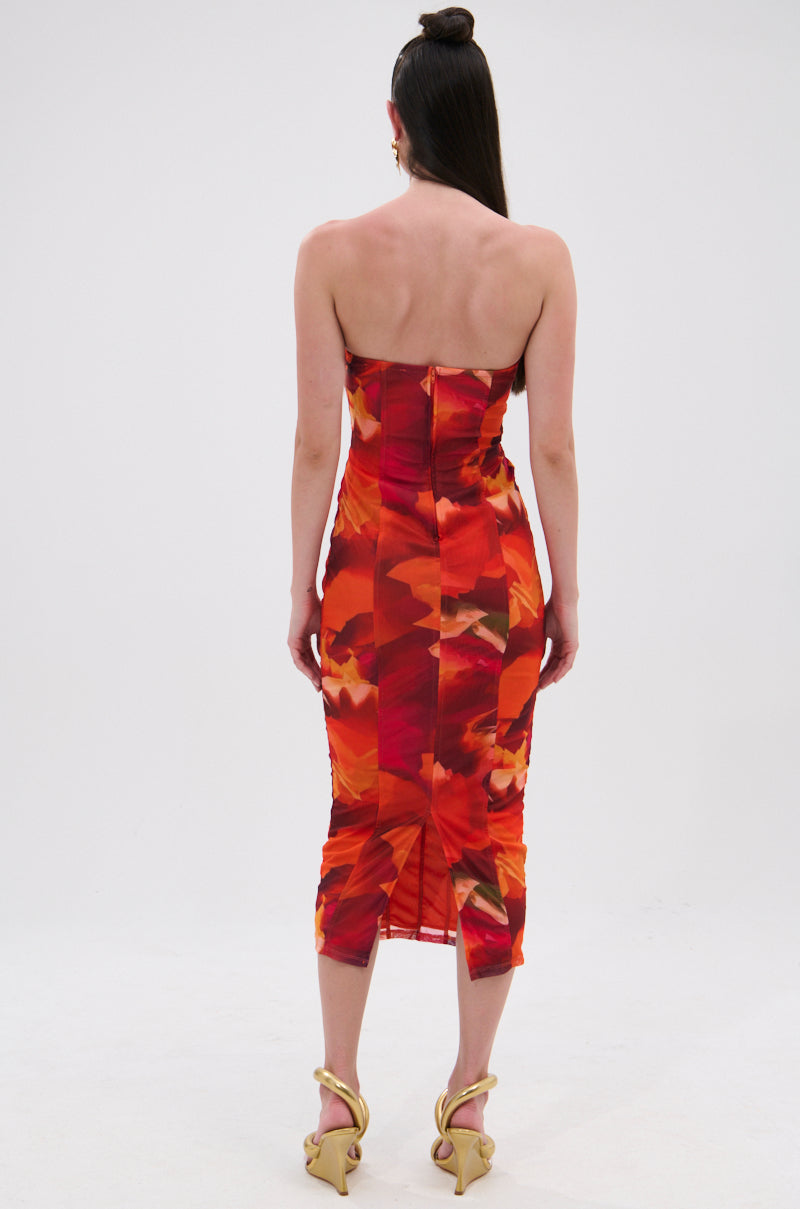 A woman with long dark hair in a half-up style faces away, modeling the ARCHETYPE PRINTED MIDI DRESS—a strapless, bodycon midi dress with a bold red and orange abstract print. She pairs it with gold high-heeled sandals against a plain white background. 4
