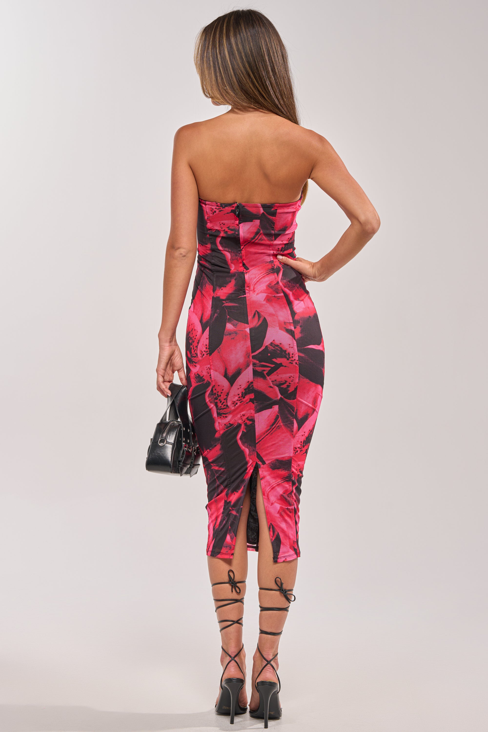 Woman stands with her back to the camera in the FRONT ROW FLORAL MIDI DRESS IN PINK MULTI, a strapless mesh dress with a body-contouring silhouette and back slit. She carries a small black handbag and wears black lace-up high-heeled sandals. 5