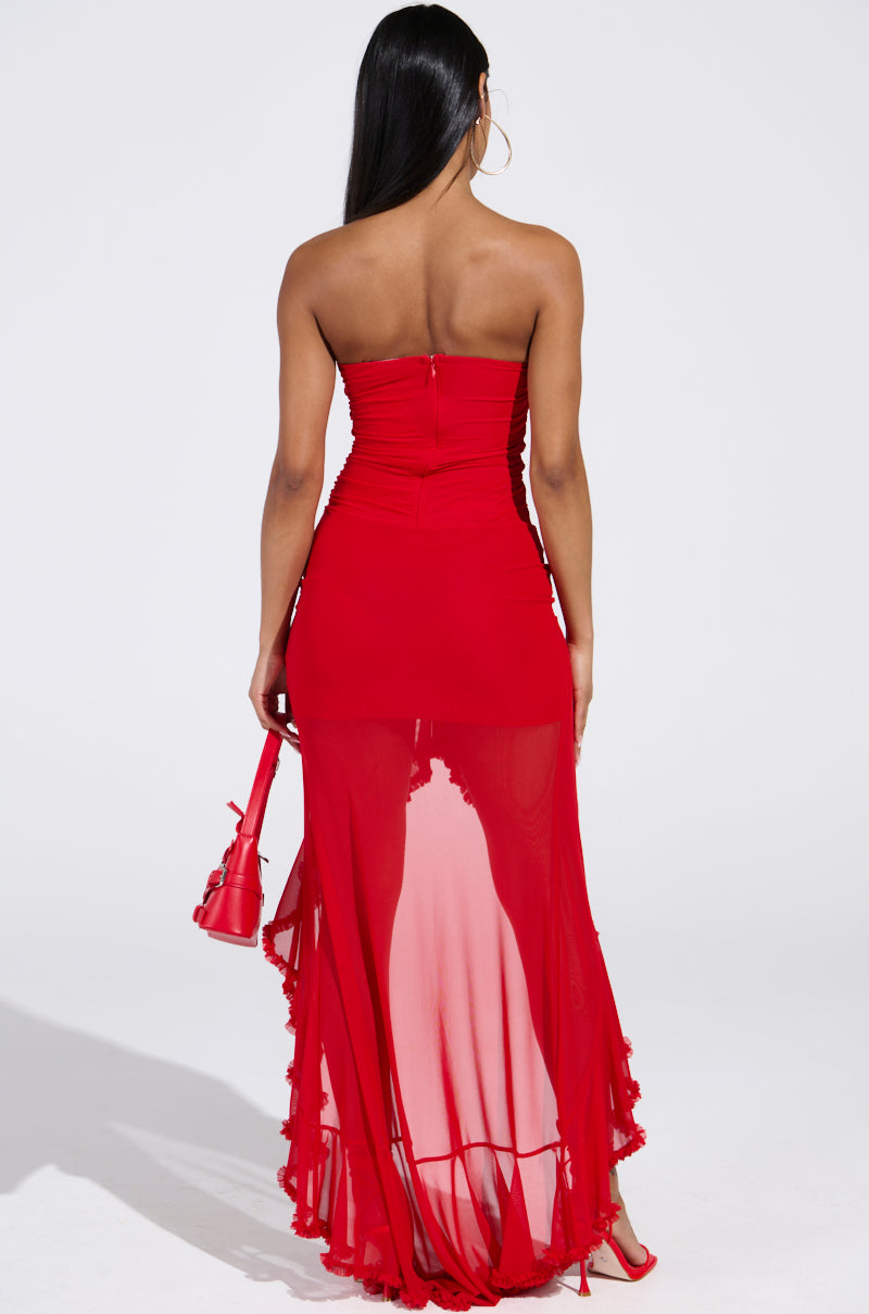A woman with long black hair, facing away against a plain white background, wears the "Oh So Hot Red Maxi Dress," featuring a strapless design with a sheer, flowing skirt and ruched detailing. She accessorizes with a small red handbag and red heels.