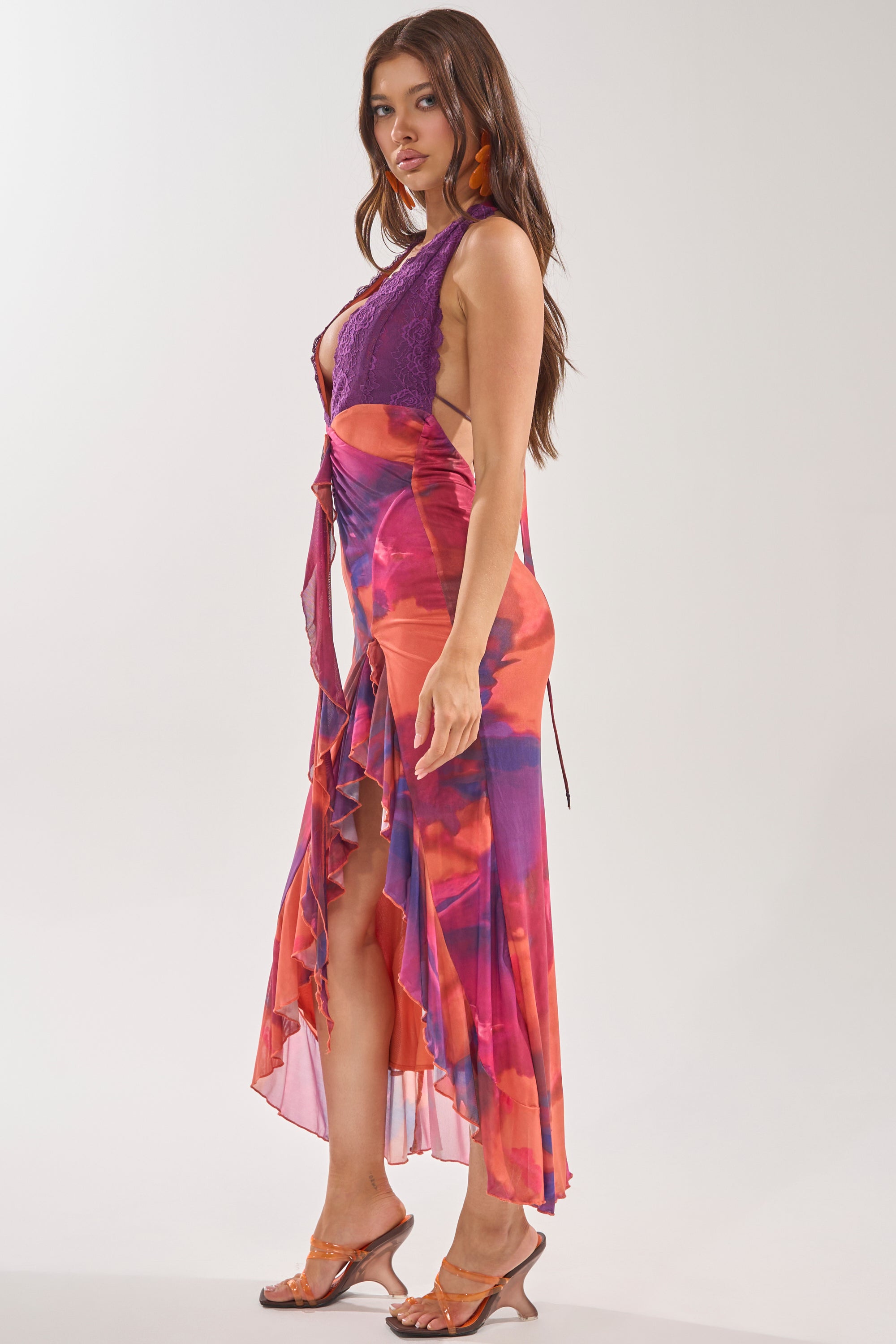 A woman with long brown hair stands sideways in the MI AMOR HALTER LACE MAXI DRESS, featuring a purple lace bodice, orange and pink sheer ruffled skirt with front slit, and clear heeled sandals against a neutral background. 2