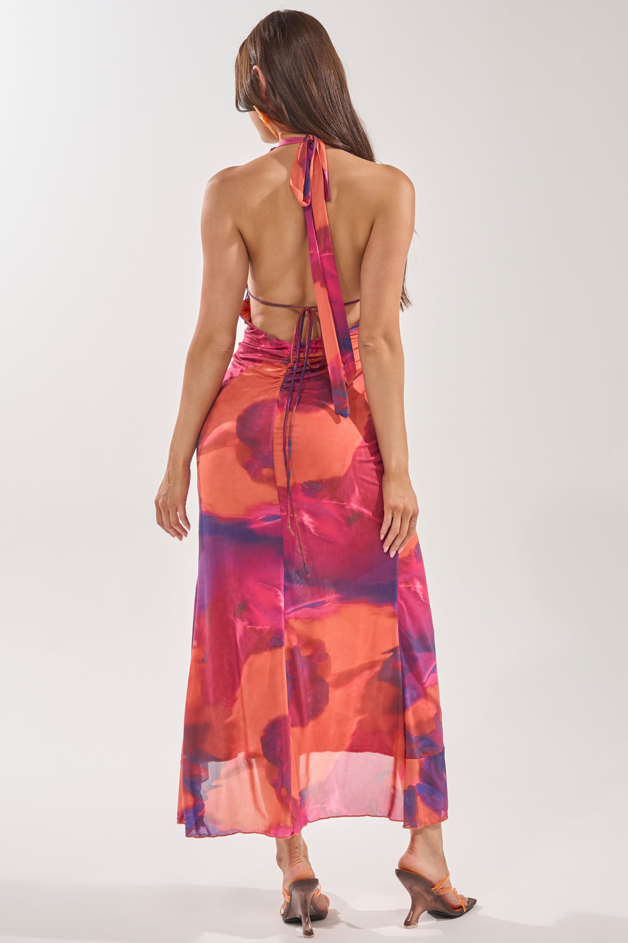 A woman with long brown hair models the MI AMOR HALTER LACE MAXI DRESS in a pink, orange, and purple abstract print with a tied neck sash. Photographed from behind, the image highlights its backless silhouette and her heeled sandals. 3