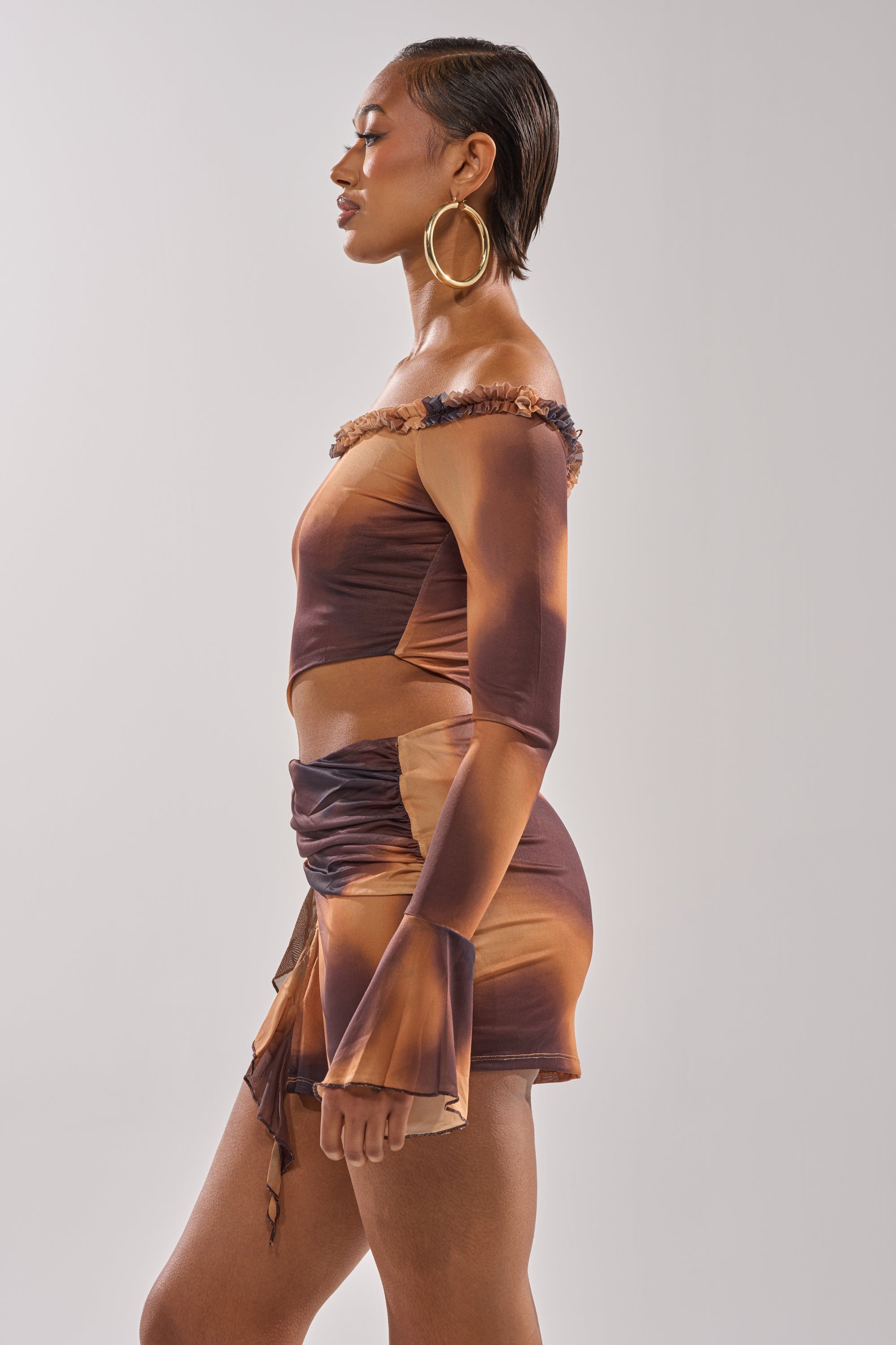 Wearing the GIVE IT TO ME BABY MINI DRESS, a person with short, slicked-back hair and large hoop earrings poses in profile against a plain light background. 2