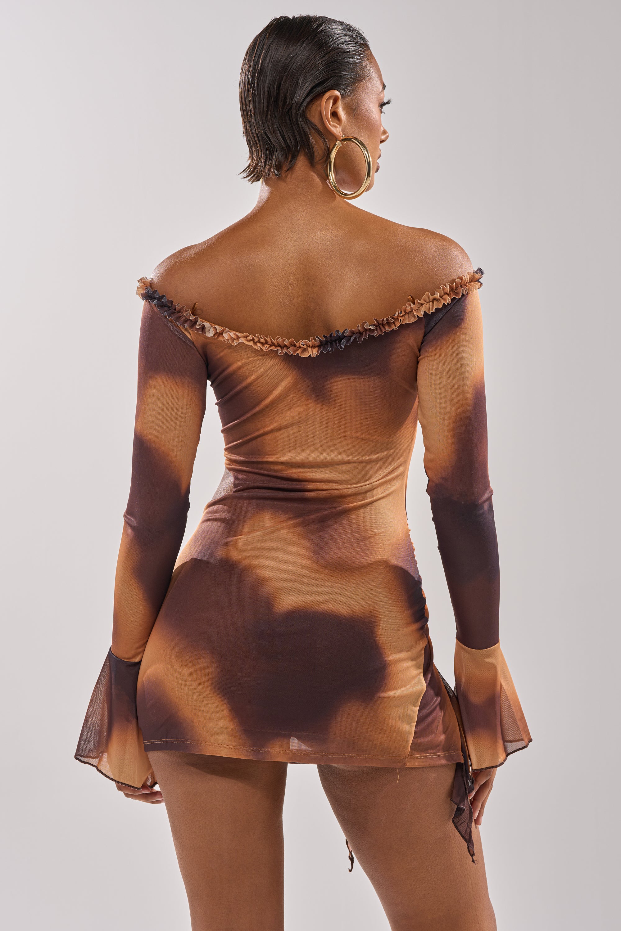 A woman with short hair and large hoop earrings faces away, wearing the GIVE IT TO ME BABY MINI DRESS—an off-shoulder, fitted mesh mini dress in a brown and tan tie-dye pattern with flared sleeves and a ruched wrap skirt. 3