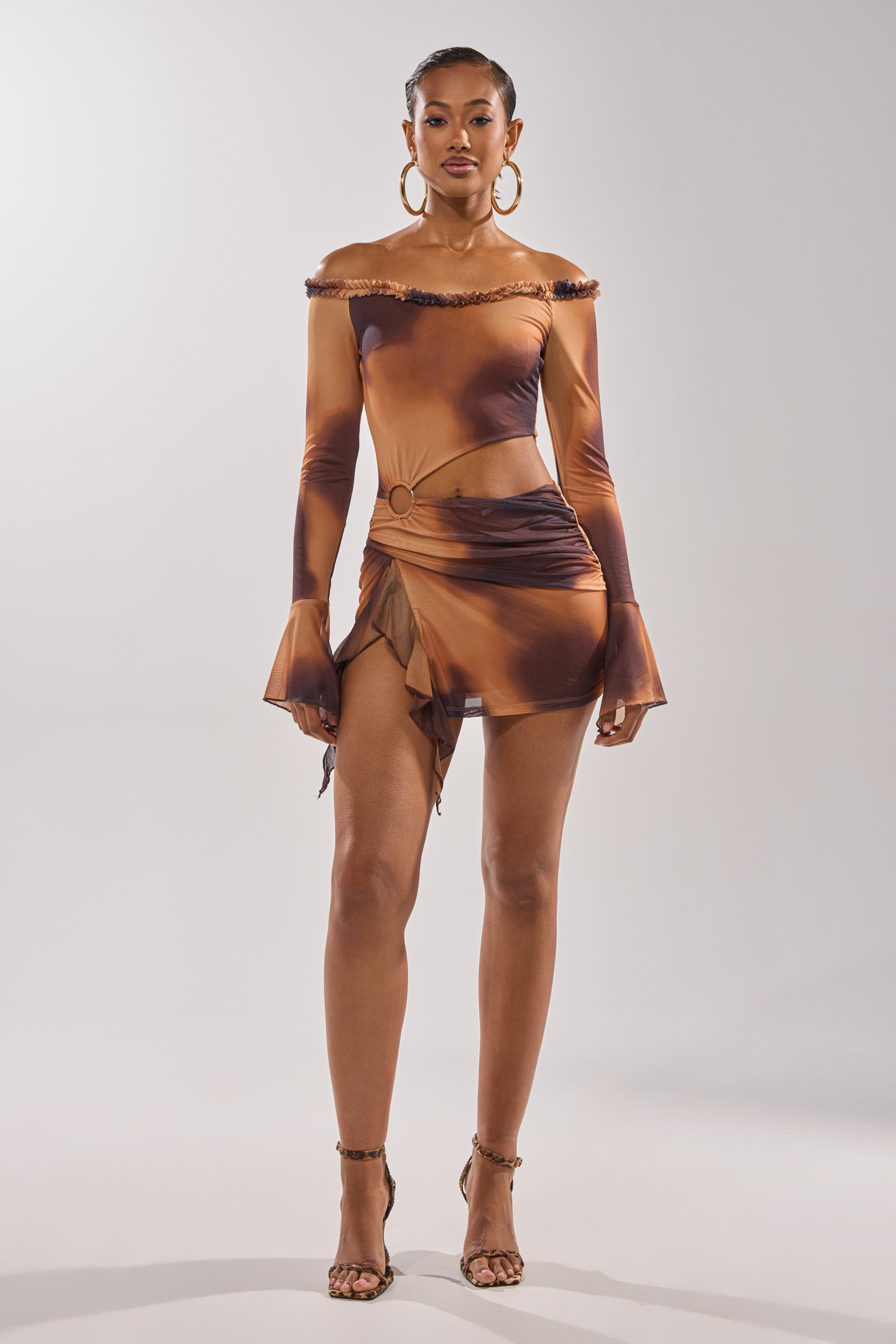 A woman exudes confidence in the GIVE IT TO ME BABY MINI DRESS—an off-shoulder brown and black mesh mini dress with long sleeves, side cutout, and ruffled hem—styled with hoop earrings and strappy high-heeled sandals. 4
