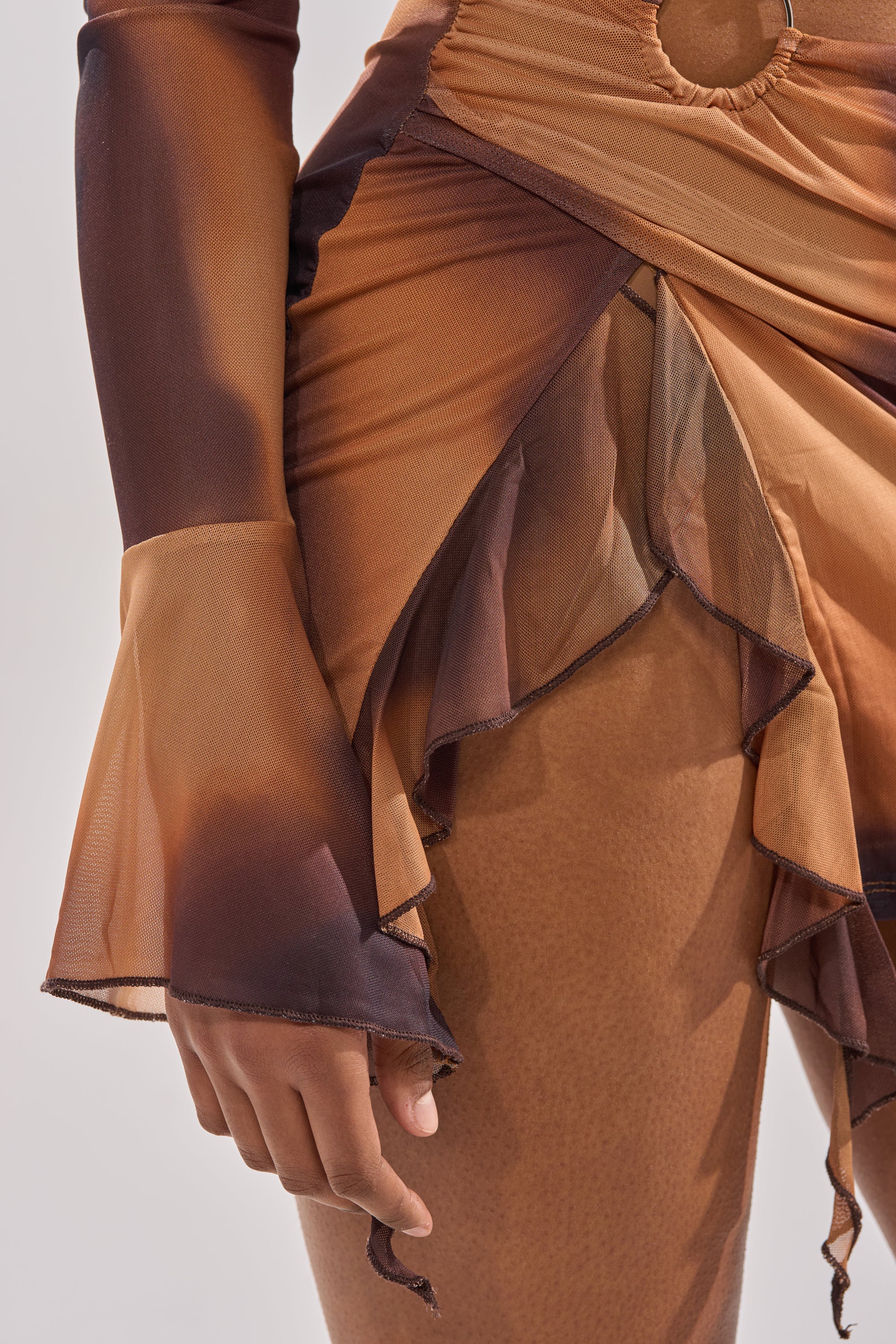 A close-up of a person wearing the GIVE IT TO ME BABY MINI DRESS, featuring sheer brown and orange ombre fabric, ruffled layers, flared sleeves, and a flowing textured look. Their hand rests relaxed by their side. 7