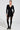 A person stands confidently in the SUSIE MINI DRESS IN BLACK, featuring a fitted design with embossed gold buttons, black tights, and pointed heels. The dress boasts long sleeves and a deep V-neck. The background is plain and well-lit.