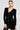 A woman wearing the Susie Mini Dress in Black, featuring an elegant bodycon silhouette, poses against a plain background. Her hair is pulled back, showcasing large hoop earrings that complement the embossed gold buttons on her attire.