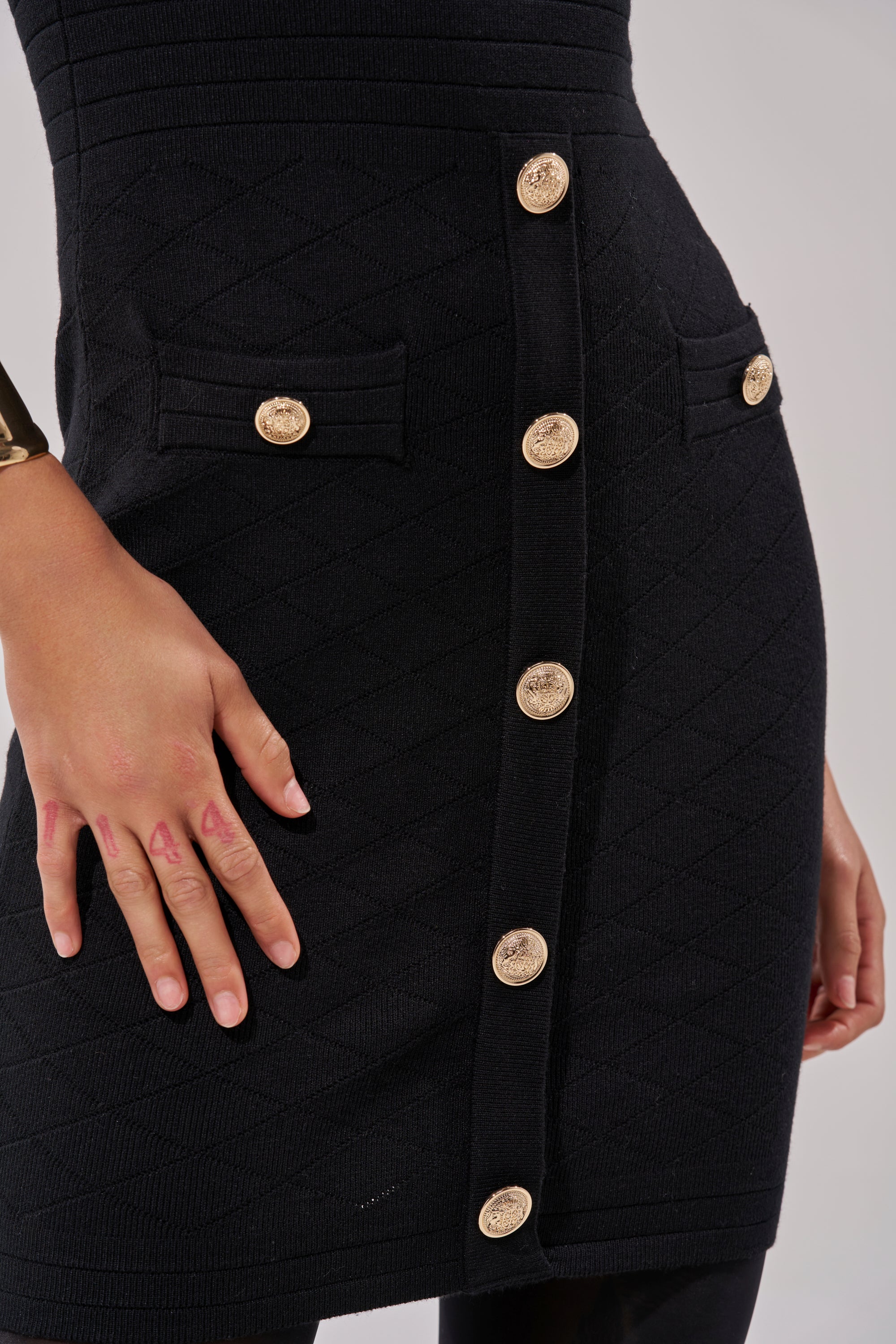 Wearing the SUSIE SLEEVELESS MINI DRESS IN BLACK, which features a fitted stretch-knit design with a diamond pattern and gold buttons, the person poses with a hand on their hip, highlighting the sculpted silhouette and a gold bracelet.