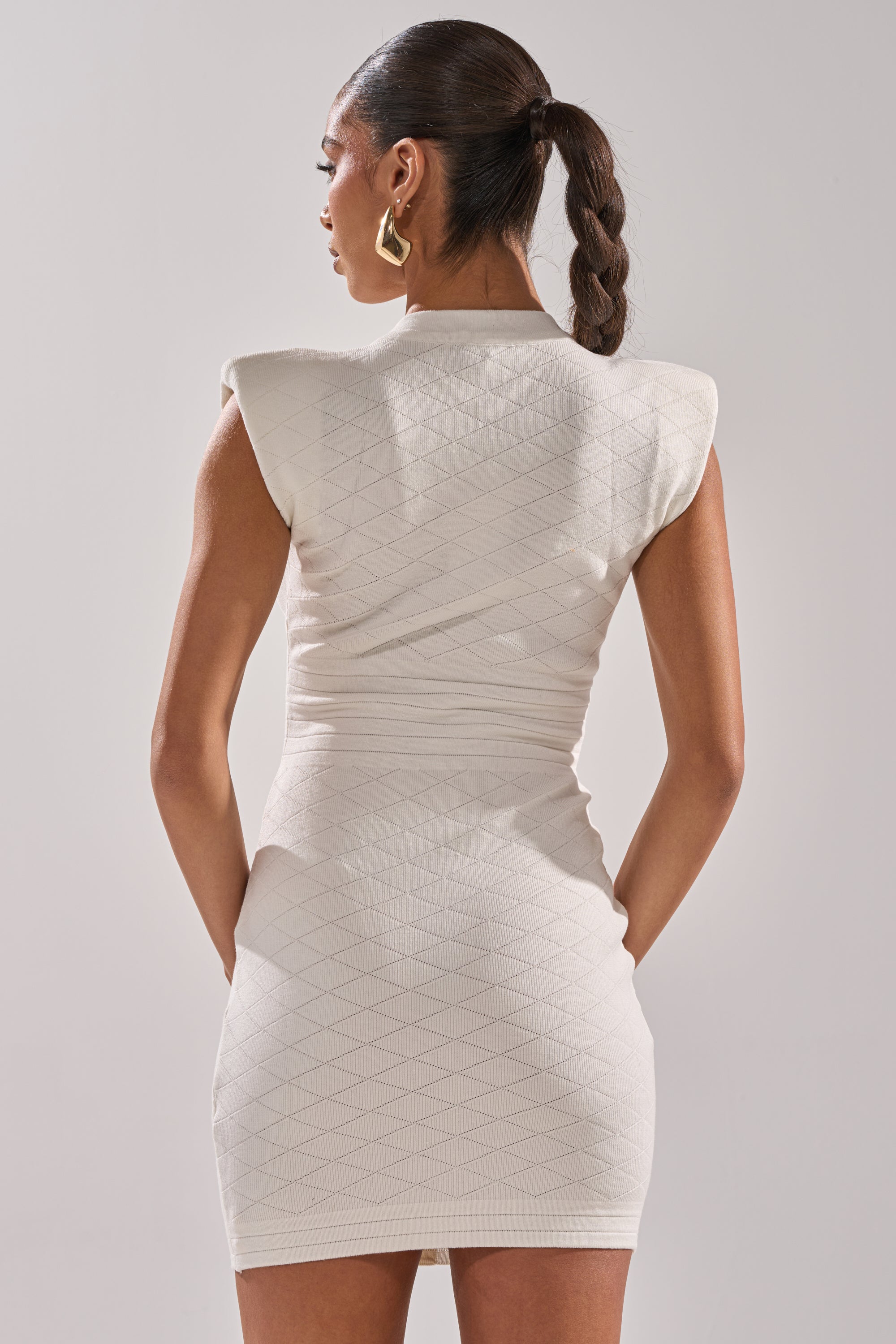 A woman with a sleek ponytail and hoop earrings is seen from behind, wearing the fitted white SUSIE SLEEVELESS MINI DRESS with textured details. Her hands rest behind her against a plain, light-colored background. 5