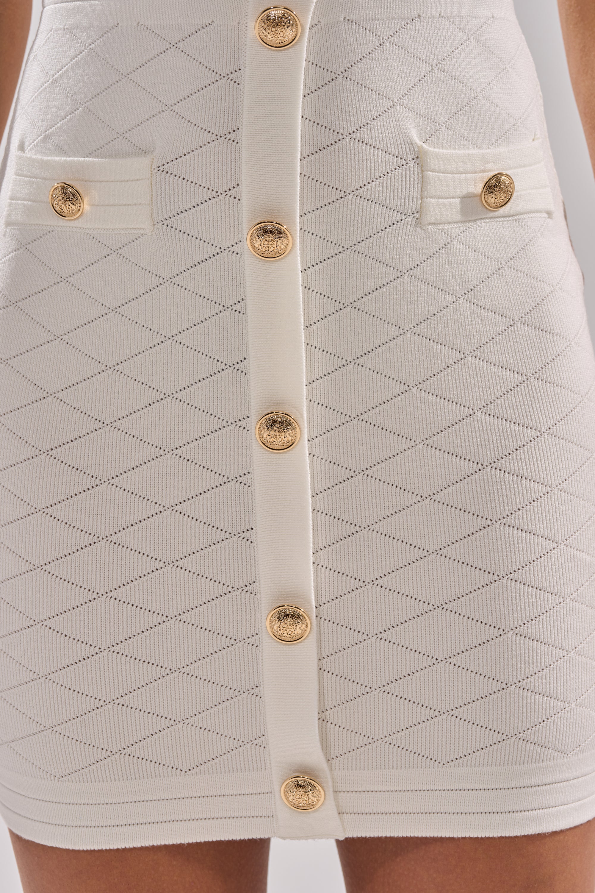 Close-up of the SUSIE SLEEVELESS MINI DRESS, a white textured stretch-knit mini dress with a diamond pattern, gold center buttons, and two small pocket flaps each adorned with a matching gold button. 7