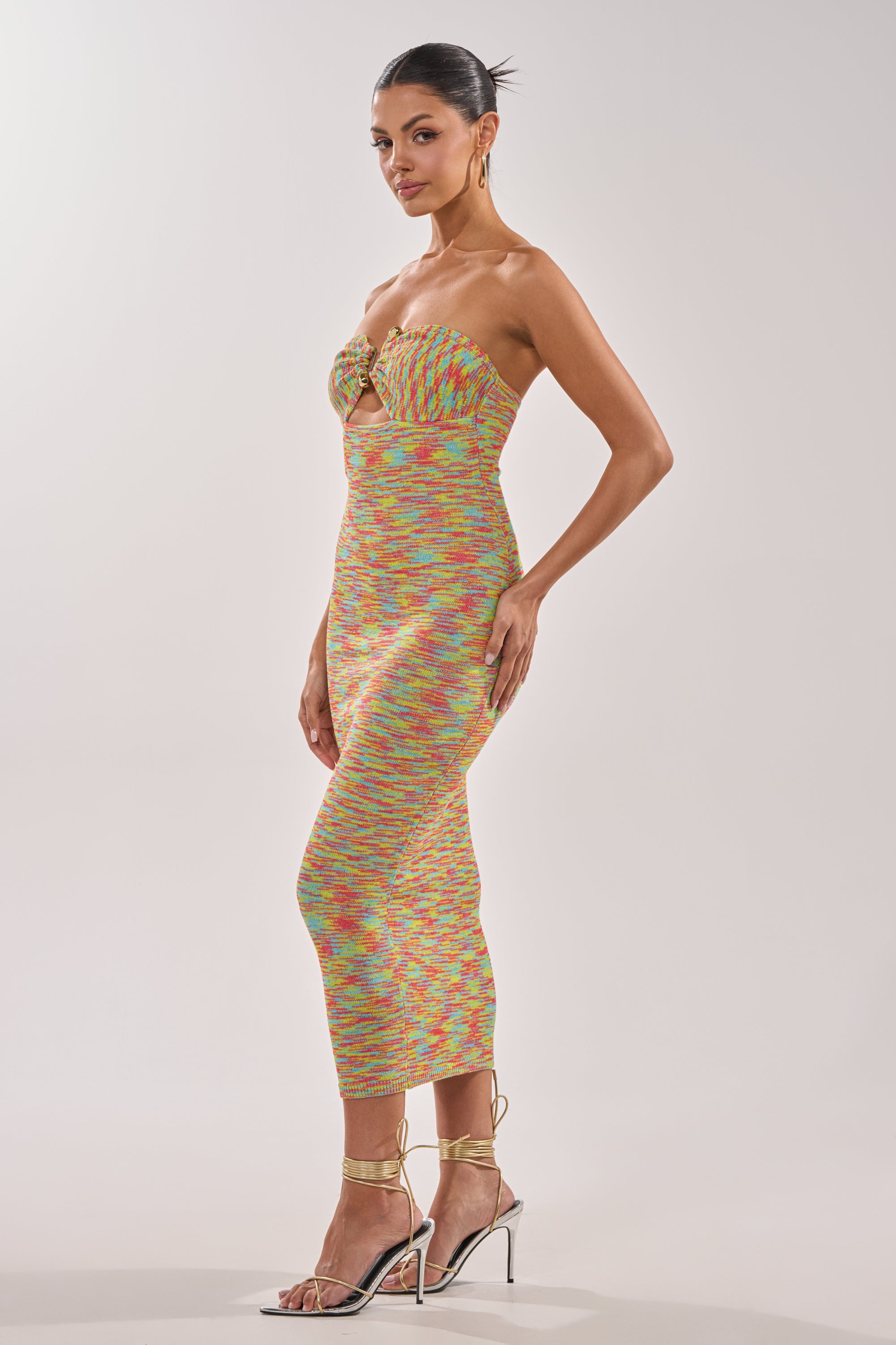 A woman models the STARRY NIGHT KNIT MIDI DRESS IN MULTI, a form-fitting, strapless dress with a front cutout and bow detail, styled with strappy gold heels, hoop earrings, and a sleek bun for a polished finish. 2