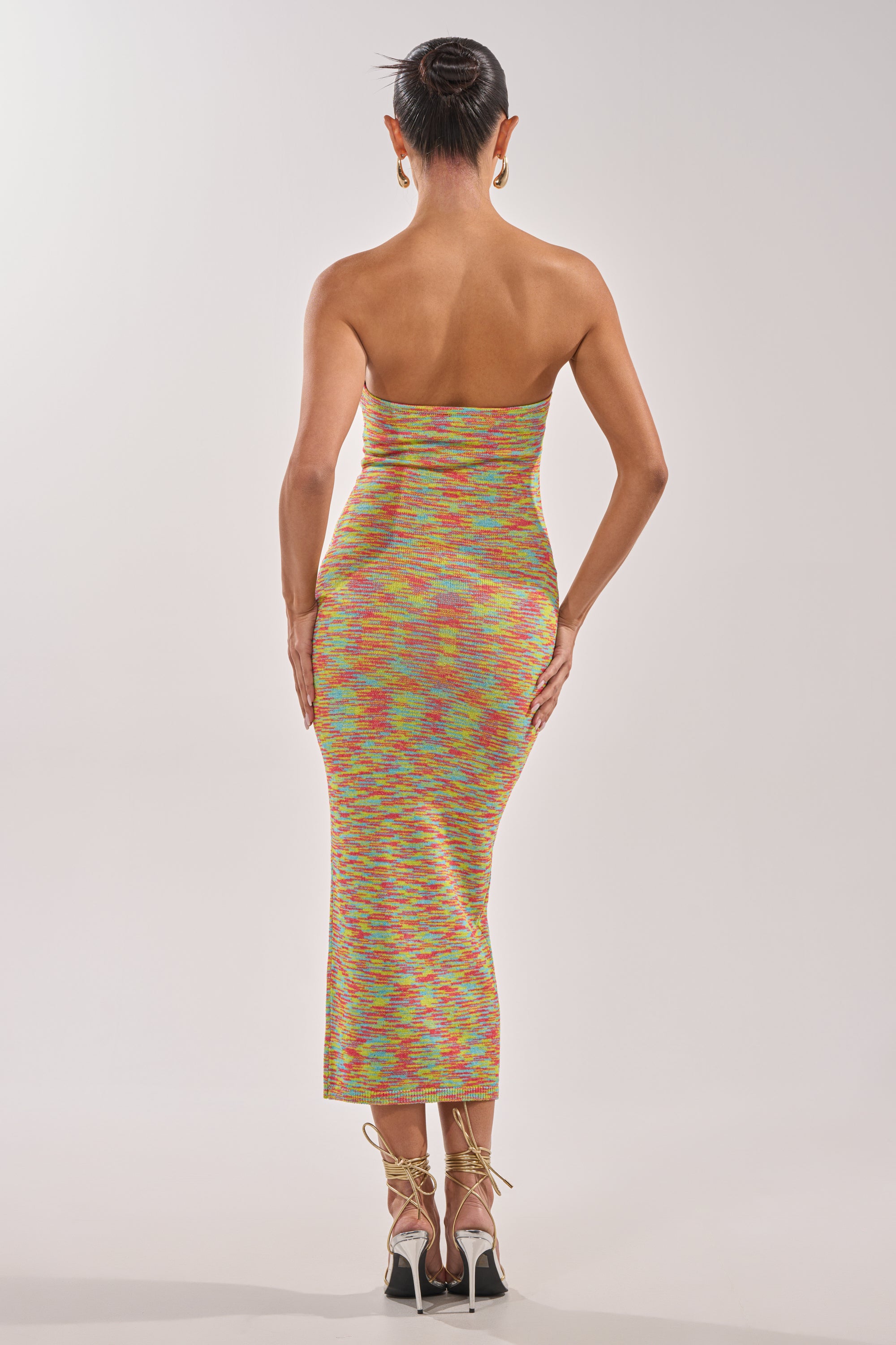 A woman stands facing away, wearing the STARRY NIGHT KNIT MIDI DRESS IN MULTI with gold hardware and high-heeled lace-up sandals. Her hair is in a neat bun as she poses against a plain light background. 3