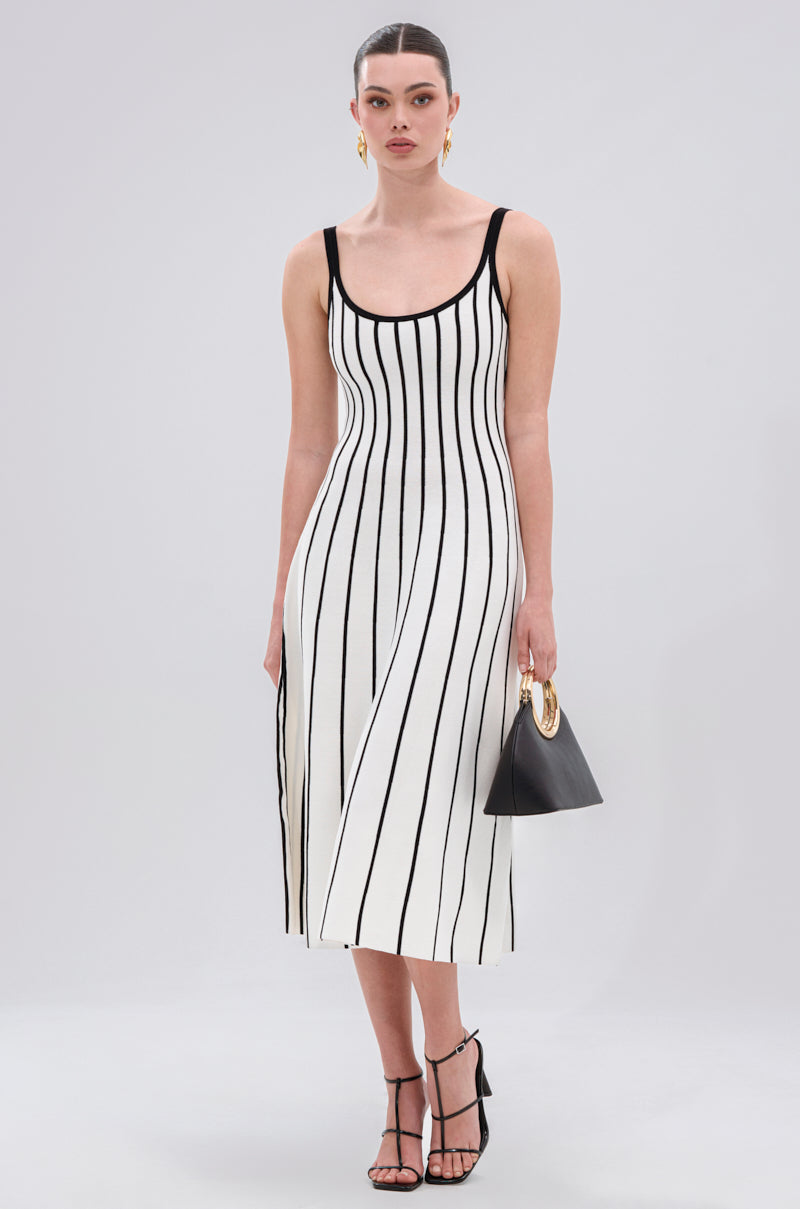 A woman models the LETS GET IT GOING KNIT STRIPE MIDI DRESS with bold black and white stripes, paired with black strappy heels, gold earrings, and a small black handbag with a gold handle against a light background.