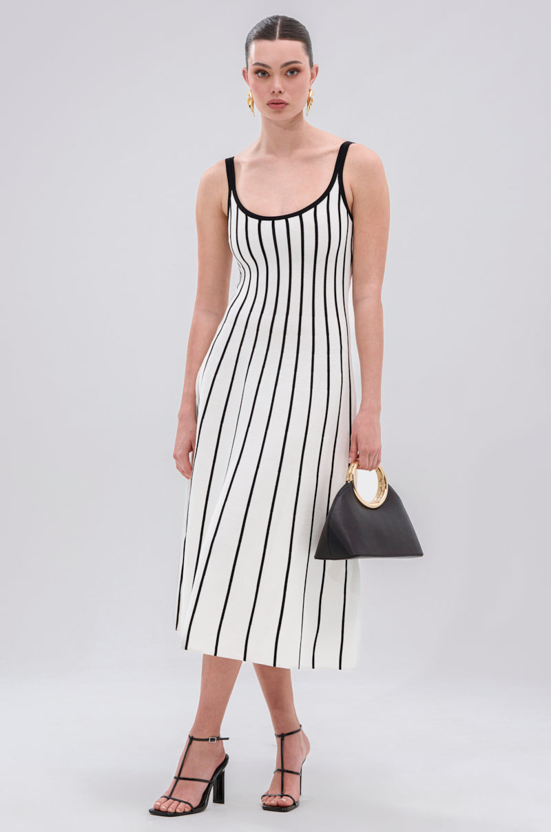 A woman wears the LETS GET IT GOING KNIT STRIPE MIDI DRESS, featuring bold black and white stripes. She accessorizes with a black handbag with a gold handle, black strappy heels, and gold earrings against a plain background.
