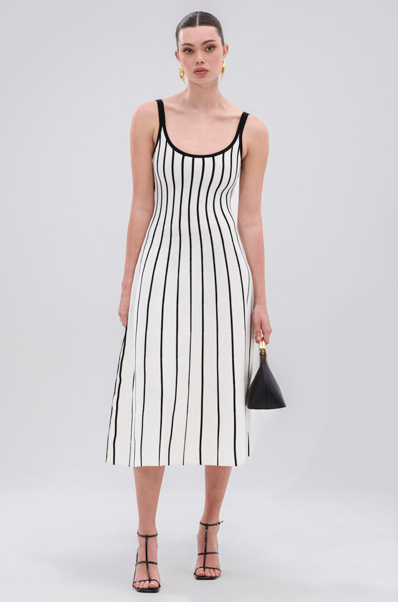 A woman wears the LETS GET IT GOING KNIT STRIPE MIDI DRESS with bold black and white stripes, paired with black heels, gold hoop earrings, and a small black handbag, standing against a light background.