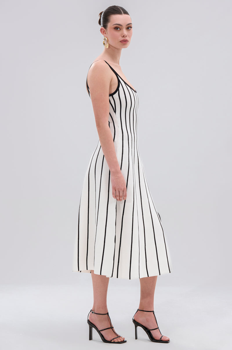 A woman stands sideways against a plain light gray background wearing the LETS GET IT GOING KNIT STRIPE MIDI DRESS, which features bold black and white stripes. She pairs it with black high-heeled sandals and large gold earrings, with her hair pulled back.