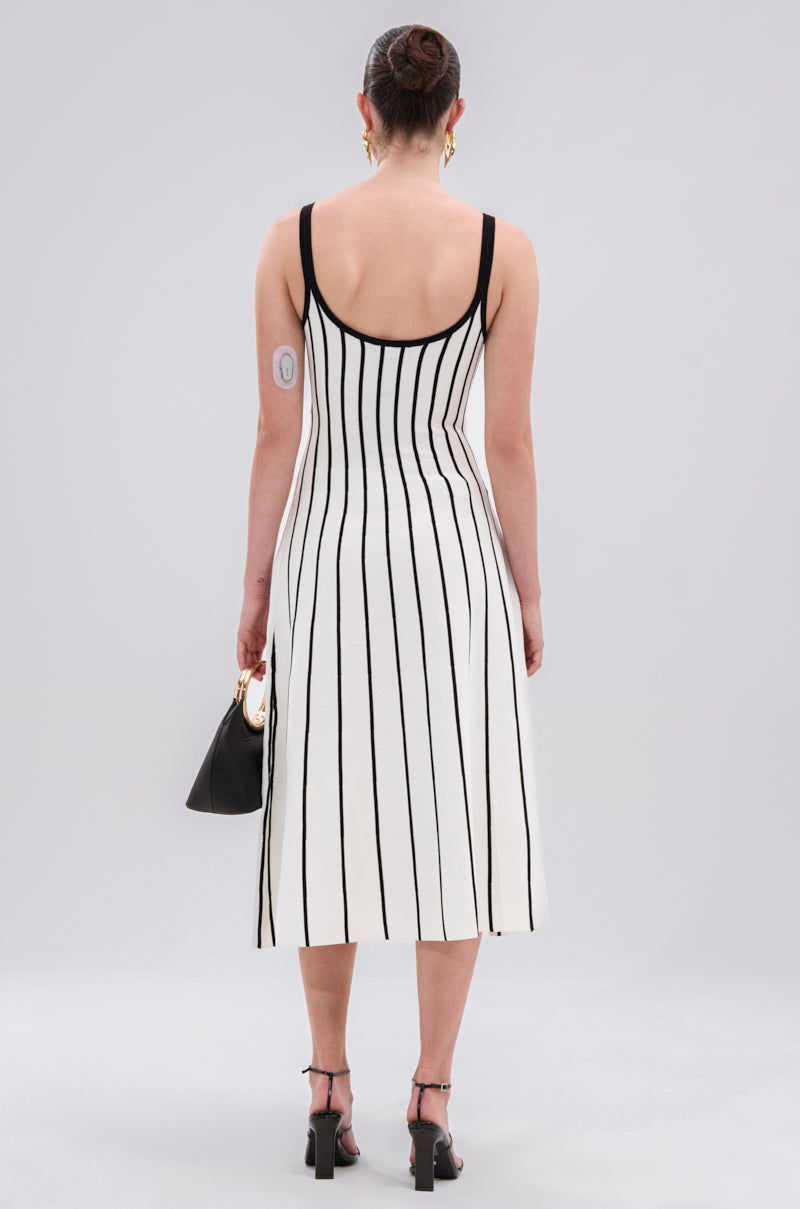 A person faces away, wearing the LETS GET IT GOING KNIT STRIPE MIDI DRESS—a bold black and white, body-hugging midi. They carry a black handbag, wear black heeled sandals, have a neat bun, and a device is visible on their upper arm.