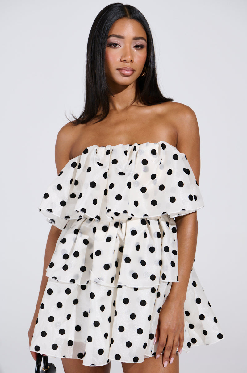 Wearing the LOVE ME LIKE THAT MINI DRESS, a woman flaunts a strapless mini adorned with black polka dots and layered ruffles, her long black hair cascading elegantly, as she clutches a small black purse against a plain white backdrop.