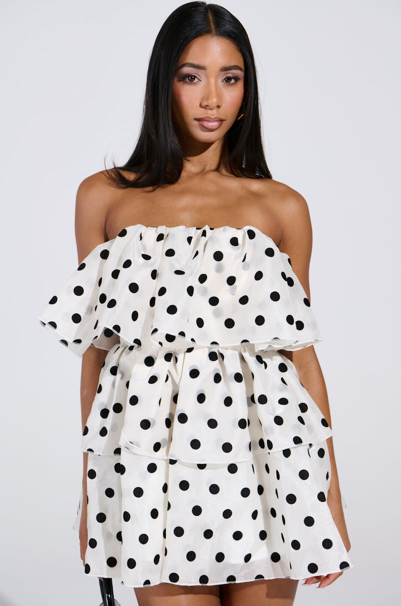 A woman with long dark hair is elegantly dressed in the LOVE ME LIKE THAT MINI DRESS, an off-the-shoulder ruffled mini dress with black polka dots. She stands against a plain white background, looking directly at the camera.