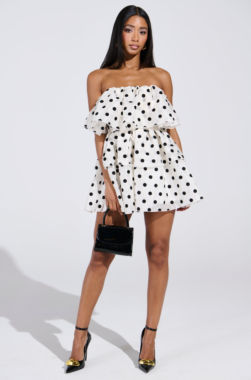 A woman exudes confidence in the LOVE ME LIKE THAT MINI DRESS, a white strapless mini adorned with black polka dots and subtle ruffles. She pairs it with a small black handbag and striking black heels with gold tips, all beautifully highlighted against the plain white background.