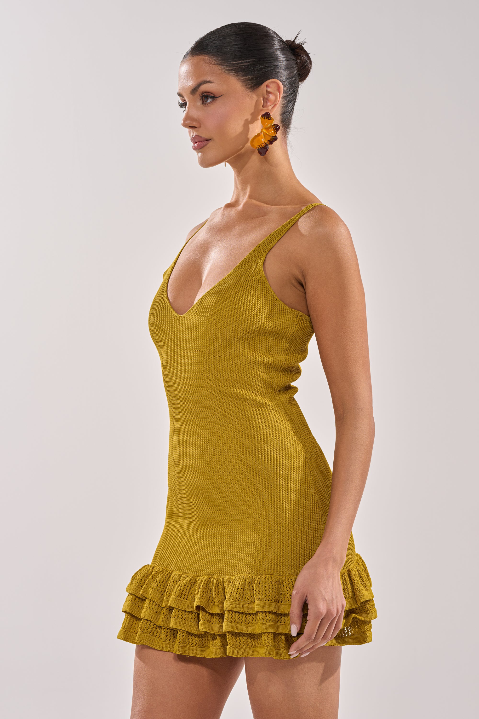 A woman in the RUFFLE QUEEN MINI DRESS IN GREEN, featuring a tank silhouette and ruffle details, stands in profile with her hair in a sleek bun and bold orange statement earrings against a light background. 2