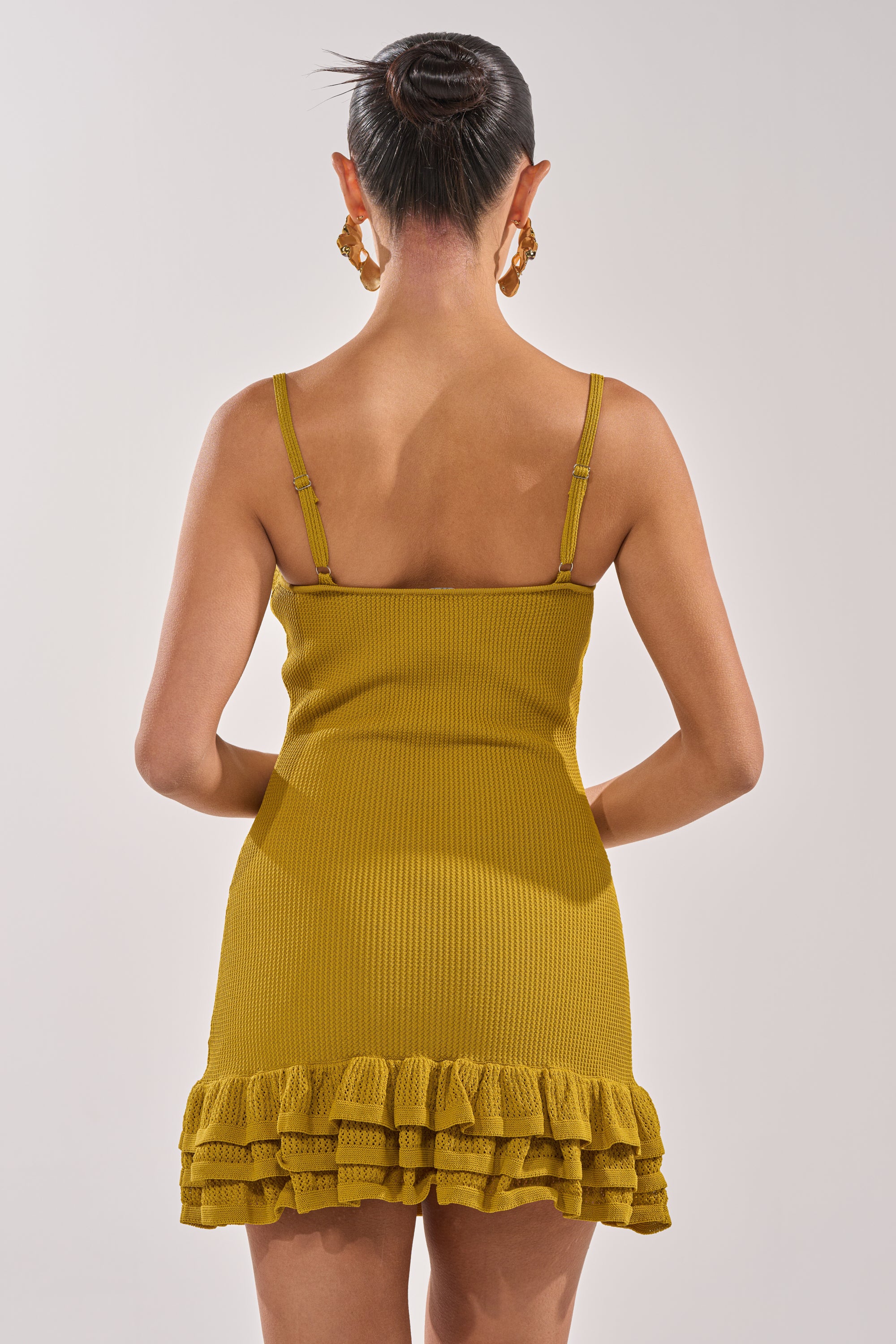 A woman with her hair in a bun and gold hoop earrings models the RUFFLE QUEEN MINI DRESS IN GREEN—featuring a chartreuse knit fabric, tank silhouette, and ruffled hem—standing with her back to the camera against a plain background. 3