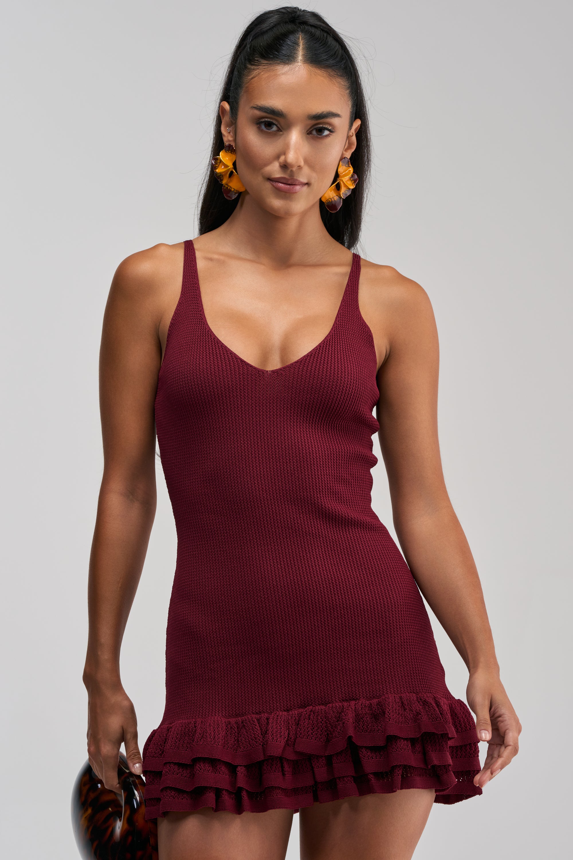 A woman wears the RUFFLE QUEEN MINI DRESS IN PLUM, featuring spaghetti straps, a V-neckline, and a ruffle hem. She accessorizes with large amber earrings and holds a tortoiseshell clutch—an effortlessly chic choice for a summer date night.