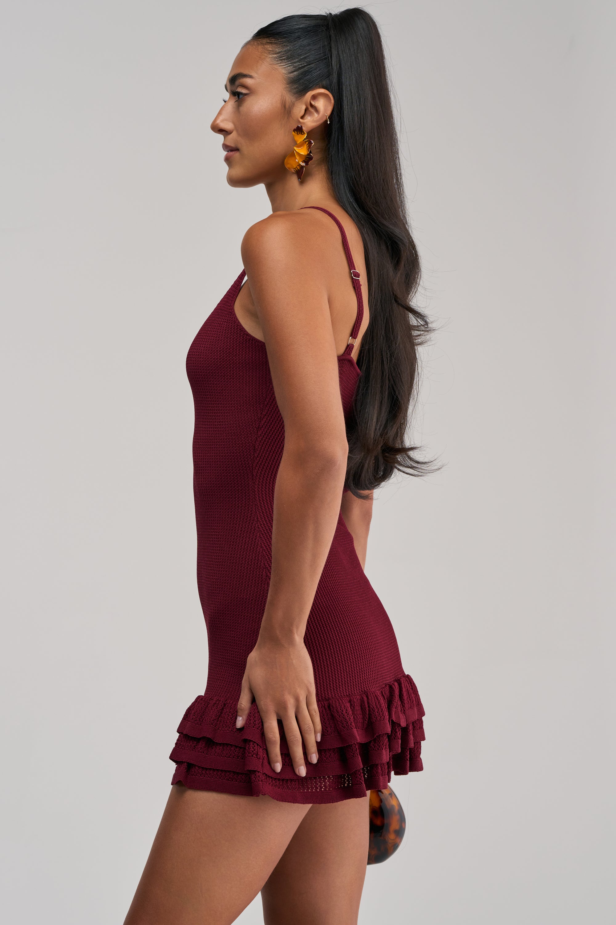Wearing the RUFFLE QUEEN MINI DRESS IN PLUM—an elegant sleeveless mini dress featuring a ribbed knit texture and playful ruffled hem—a woman stands in profile against a plain light backdrop. Her long dark hair is pulled into a ponytail, accented by bold orange statement earrings—an effortlessly chic summer date night look.