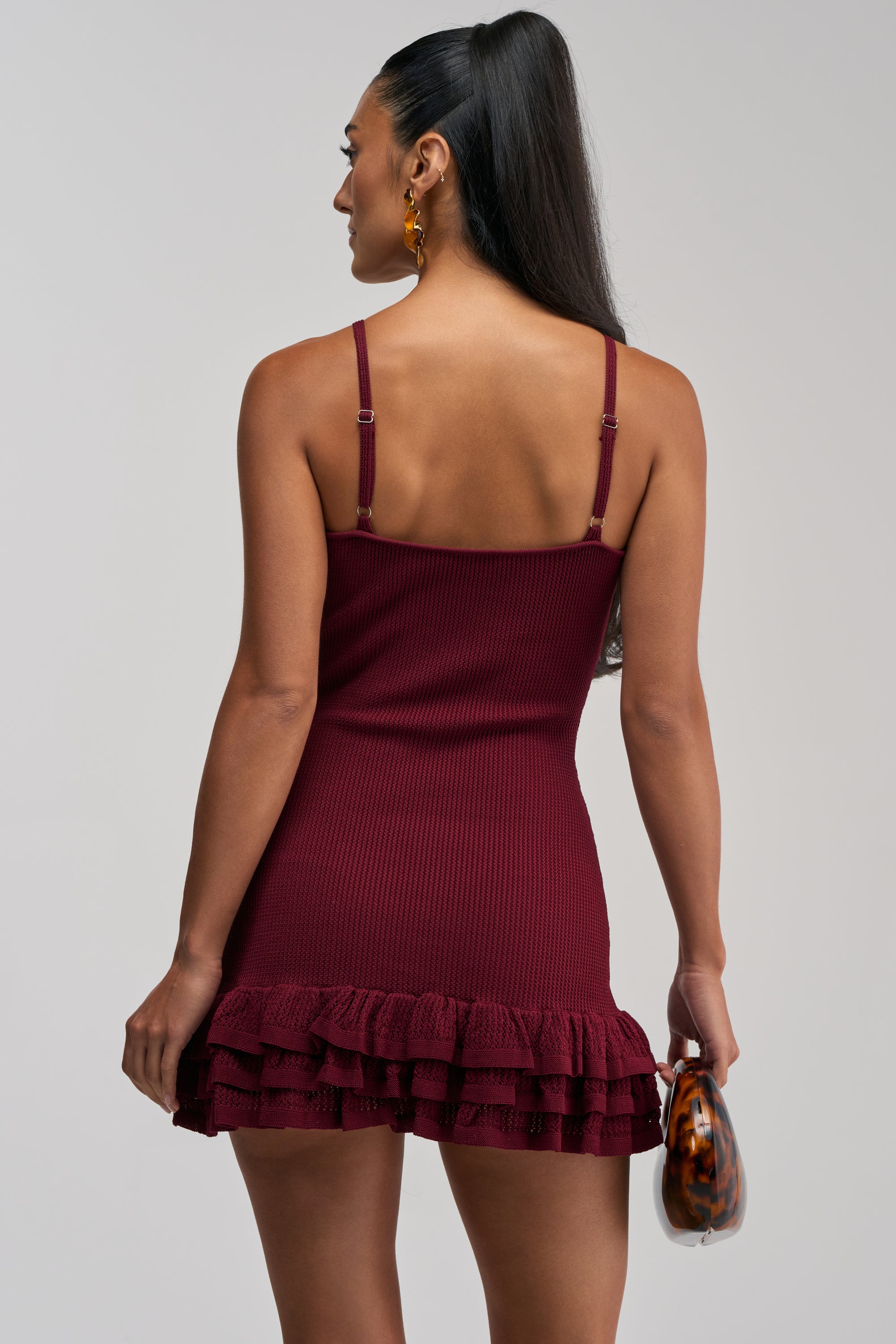 A woman with long dark hair styled in a ponytail wears the RUFFLE QUEEN MINI DRESS IN PLUM—a sleeveless, ribbed knit mini dress featuring a ruffled hem that’s ideal for summer date nights. Holding a brown patterned handbag, she faces away from the camera against a plain background.