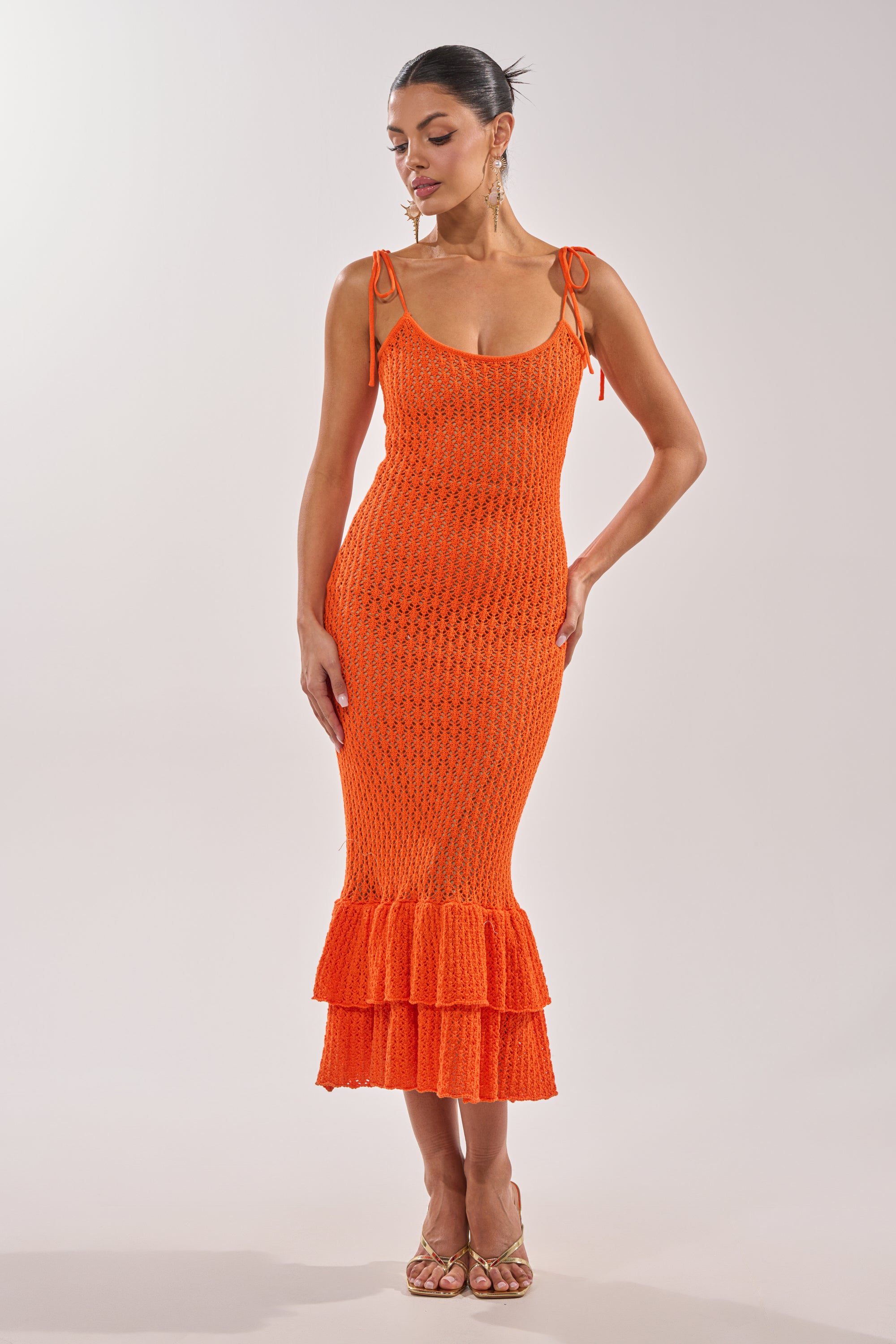 A woman models the LINSEY MAXI KNIT DRESS IN ORANGE—a sleeveless maxi dress with shoulder ties and a ruffled hem—paired with strappy gold heels, set against a plain light backdrop. 1