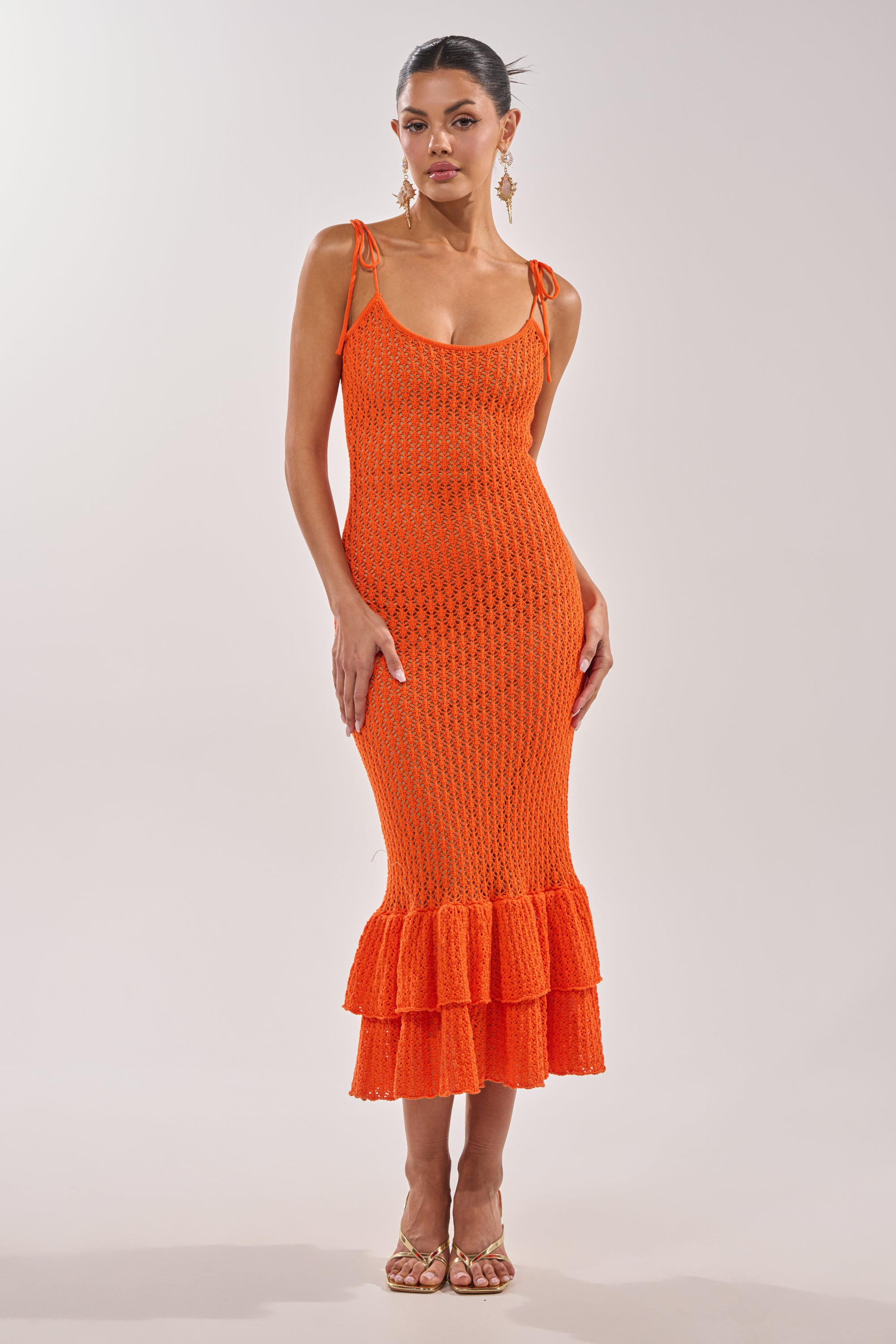 A woman wears the LINSEY MAXI KNIT DRESS IN ORANGE, featuring sleeveless shoulder tie straps and a ruffled hem, styled with gold sandals and statement earrings, against a plain light background. 2