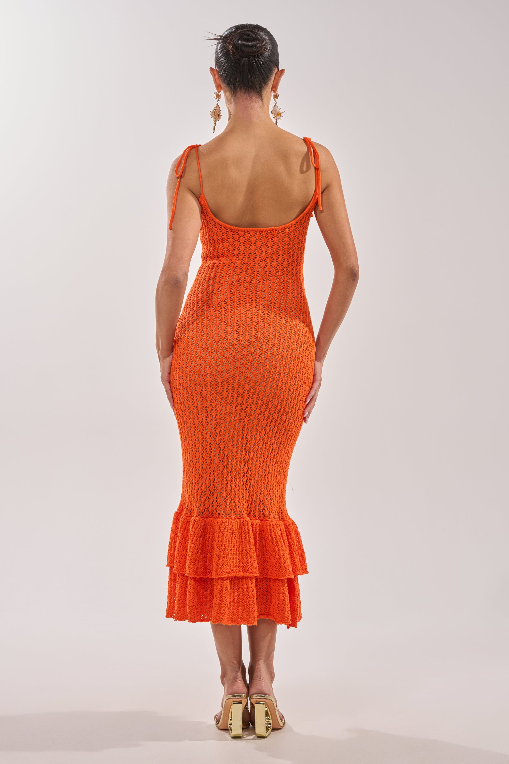 A woman with her hair in a bun and large earrings faces away, wearing the LINSEY MAXI KNIT DRESS IN ORANGE—an orange sleeveless knit maxi with shoulder ties and a ruffled hem—styled with flat sandals. 4